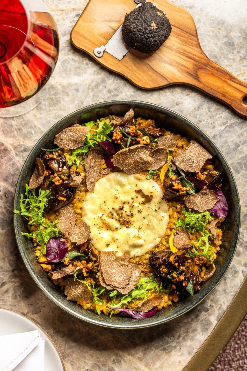 A bowl of risotto topped with soft cheese, garnished with fresh greens, shaved truffles, and black and yellow turnip. Accompanying the dish is a glass of red wine and a black truffle on a wooden cutting board.