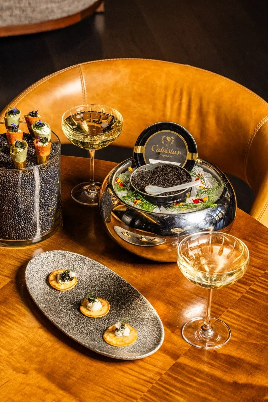 A table set with two glasses of champagne, a small oval dish with three bites of caviar on crackers, a bowl of Calvisius caviar with a spoon, a container of black caviar topped with edible flowers and greens, and a glass bowl of canapé bites topped w