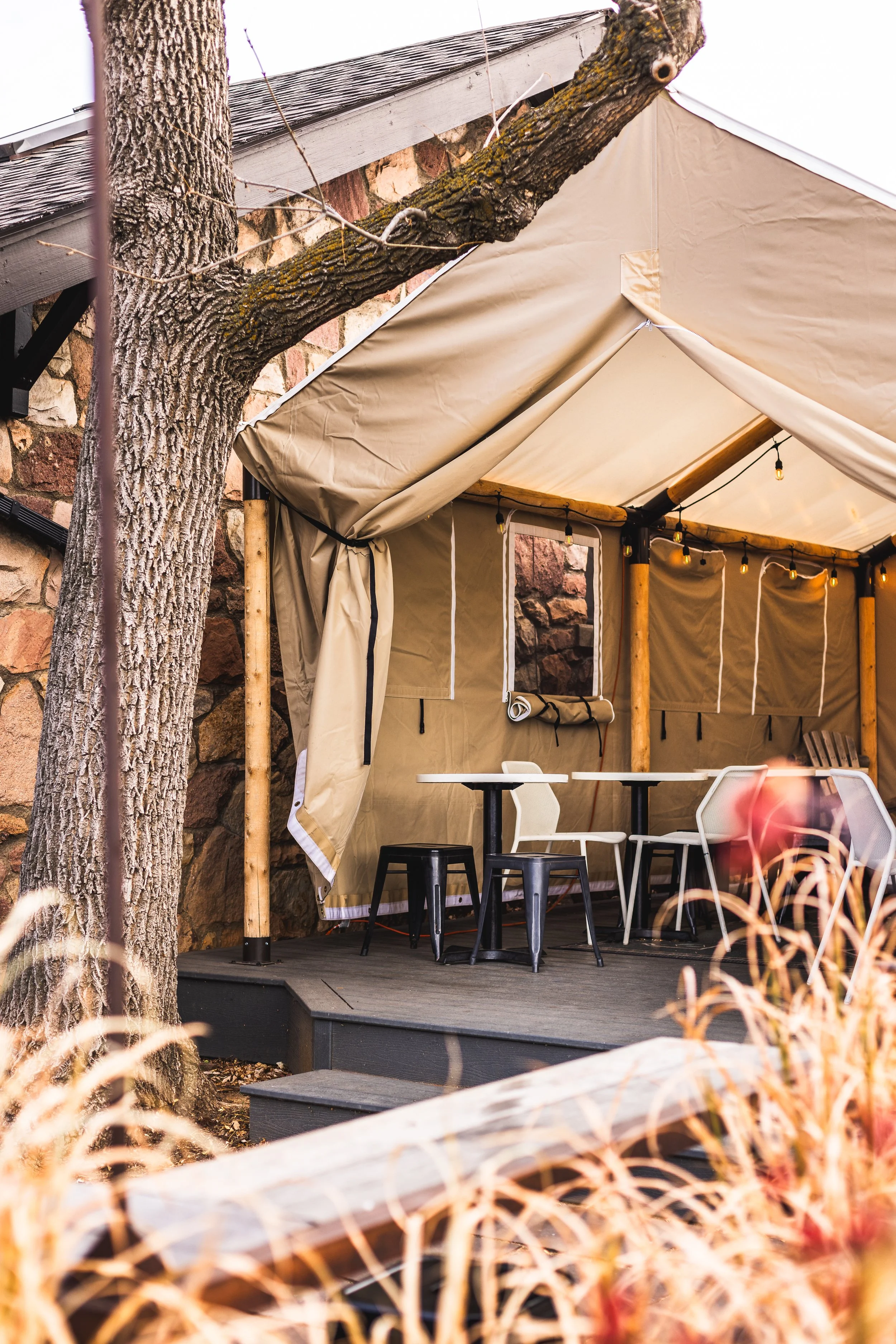 An outdoor tented seating area with a small table and various chairs, situated on a wooden platform, surrounded by a tree and some grass and plants.