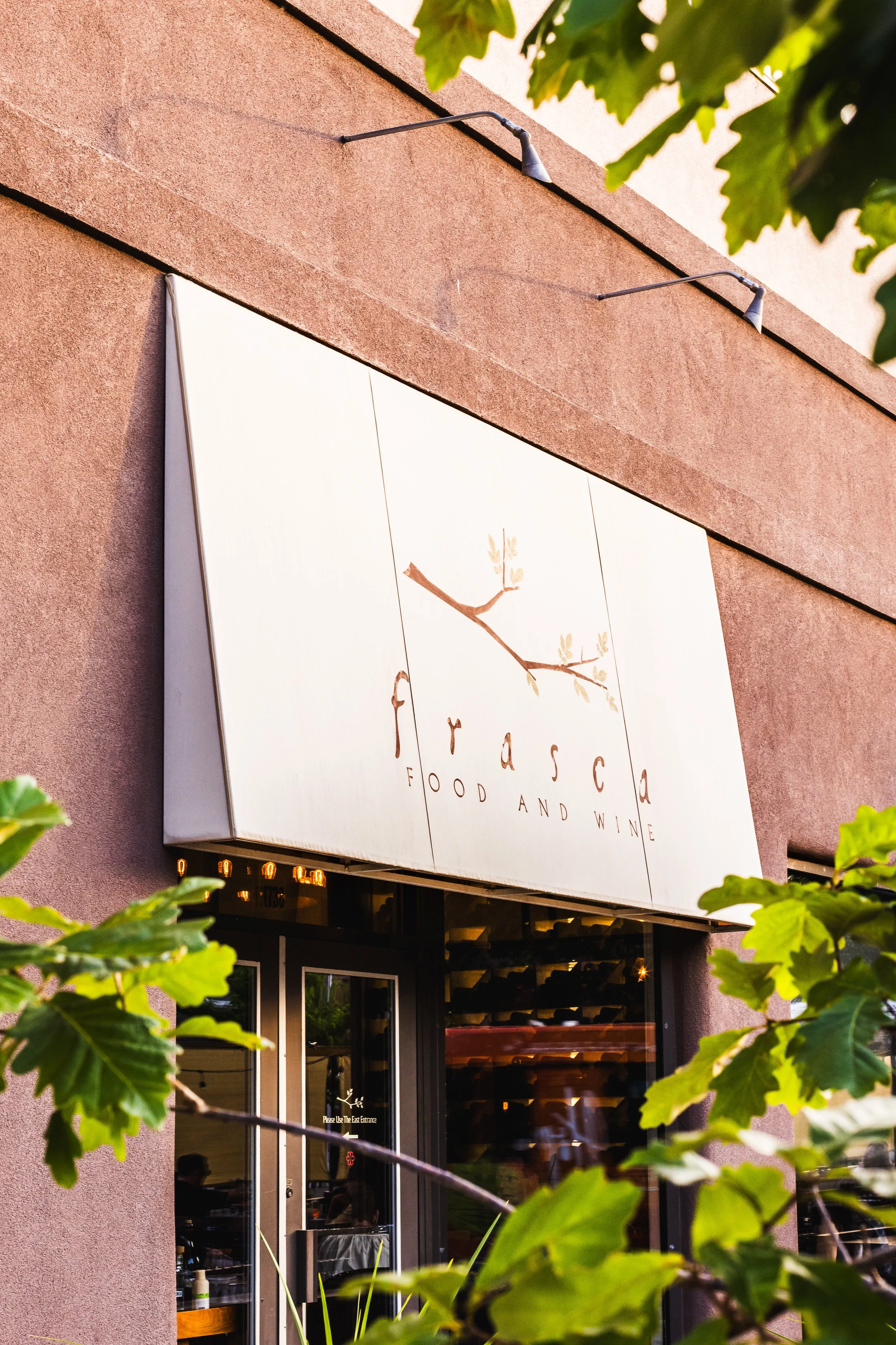 Exterior view of Frasca Food and Wine restaurant with a white sign featuring a branch and leaves above the entrance, partially obscured by green leaves.