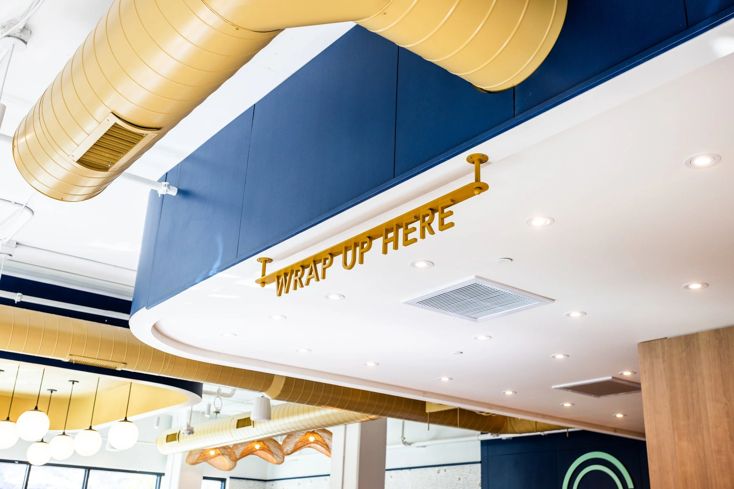 Interior of a modern space with a prominent yellow duct, blue and white ceiling, sign that reads 'Wrap Up Here' hanging from the ceiling, and ceiling lights.