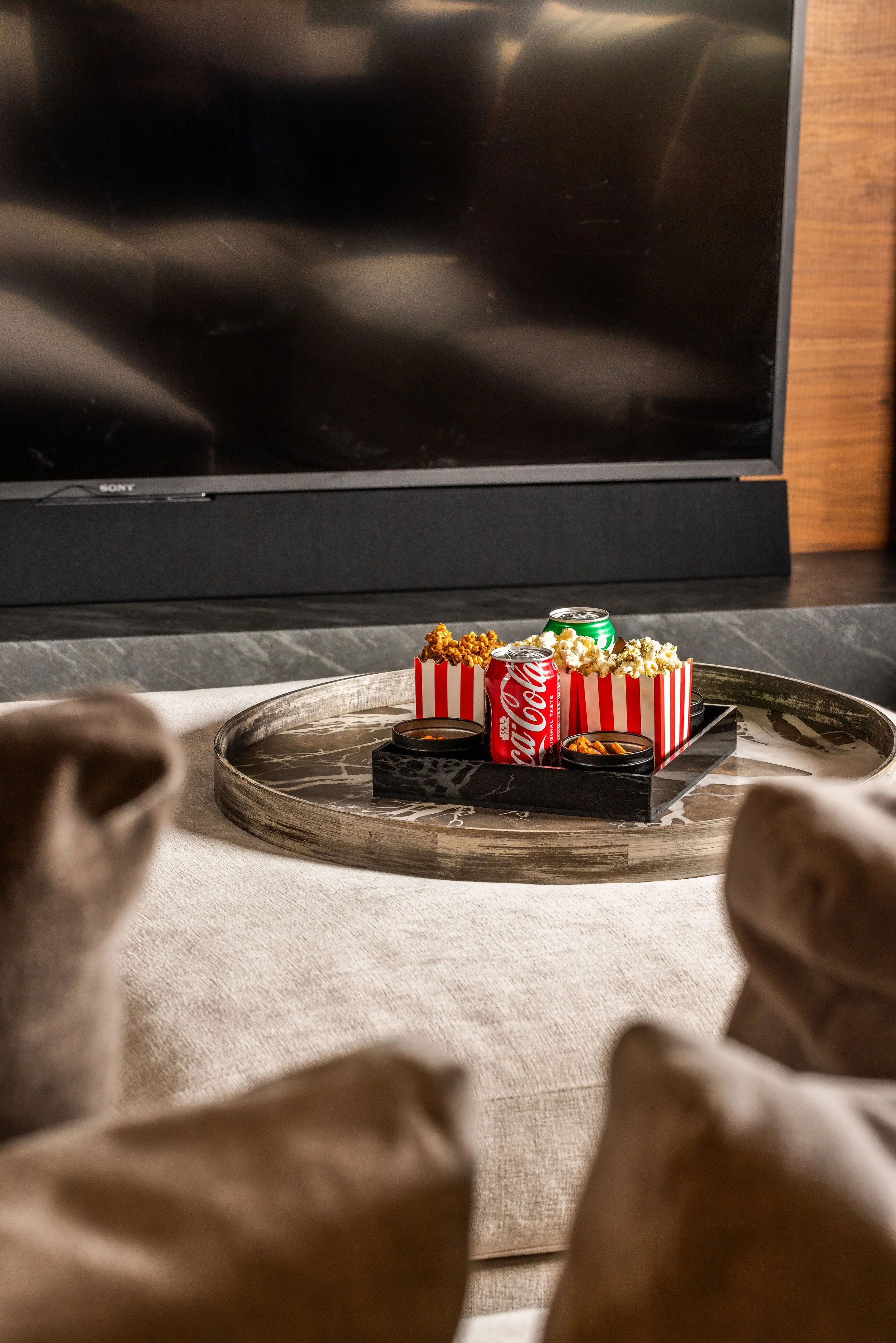 A tray with popcorn, two cans of soda, and small containers of snacks on a coffee table in front of a TV.