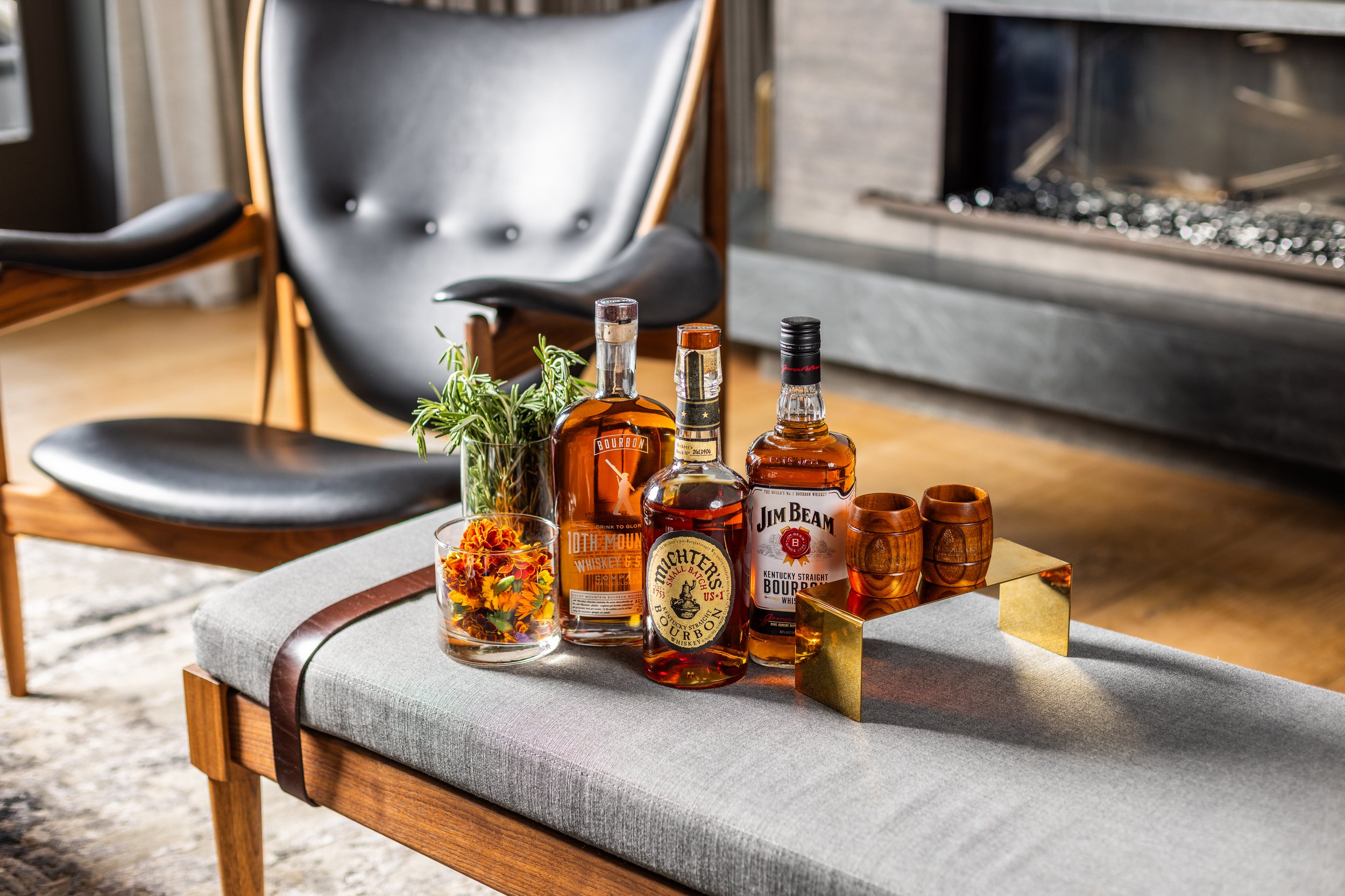 A coffee table with bottles of bourbon whiskey, wooden cups, a plant, and a small container of flowers, with a black chair and a fireplace in the background.
