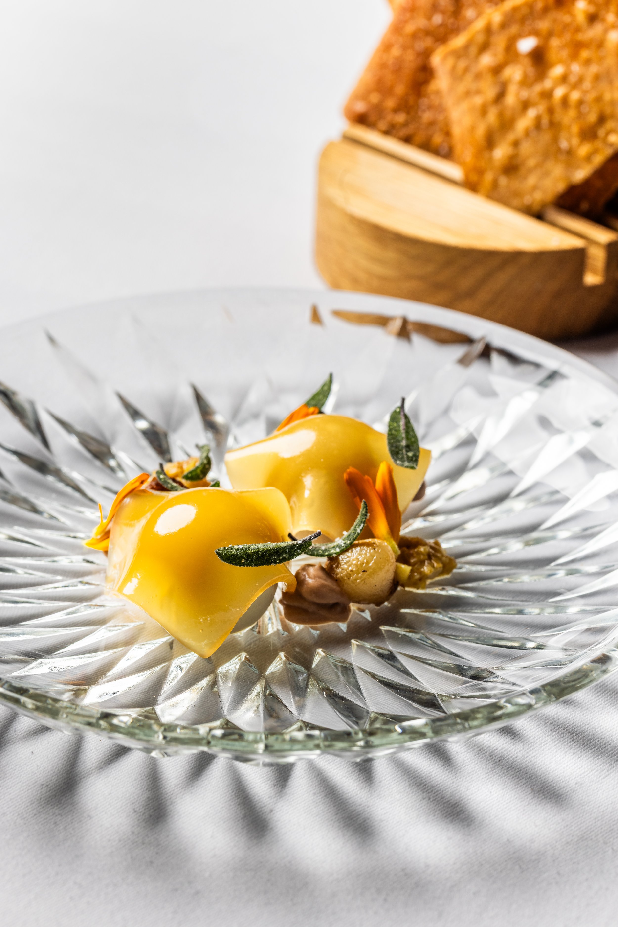 Elegant dessert with two yellow glazed pastries garnished with small green leaves on a decorative glass plate, with a wooden container of rectangular brown bread slices in the background.