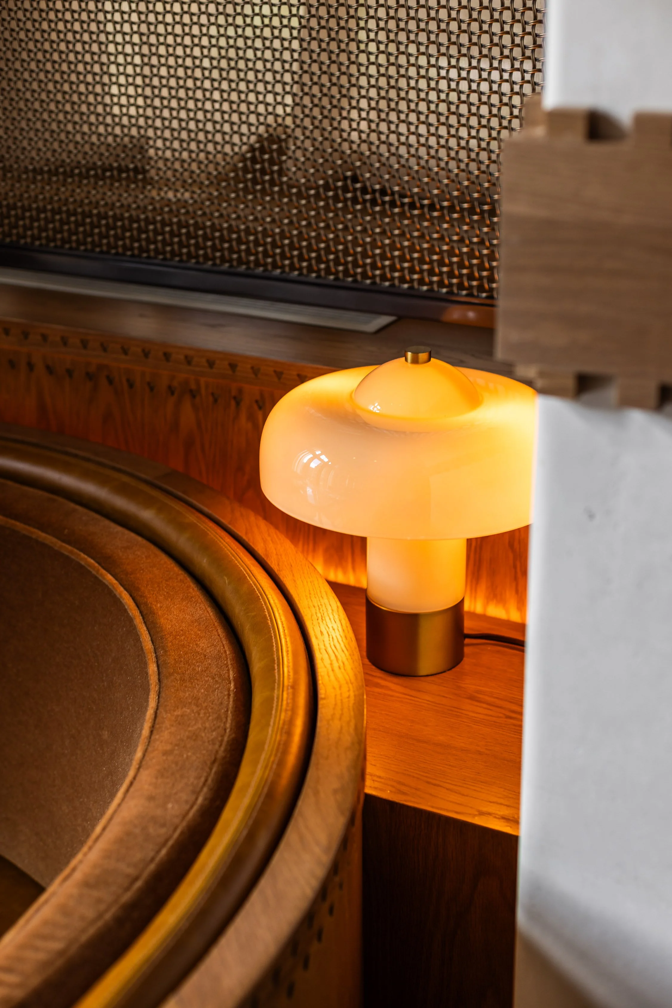 A modern table lamp with a glossy, milky glass shade and a black base on a wooden side table next to a curved brown upholstered chair in a warmly lit room.