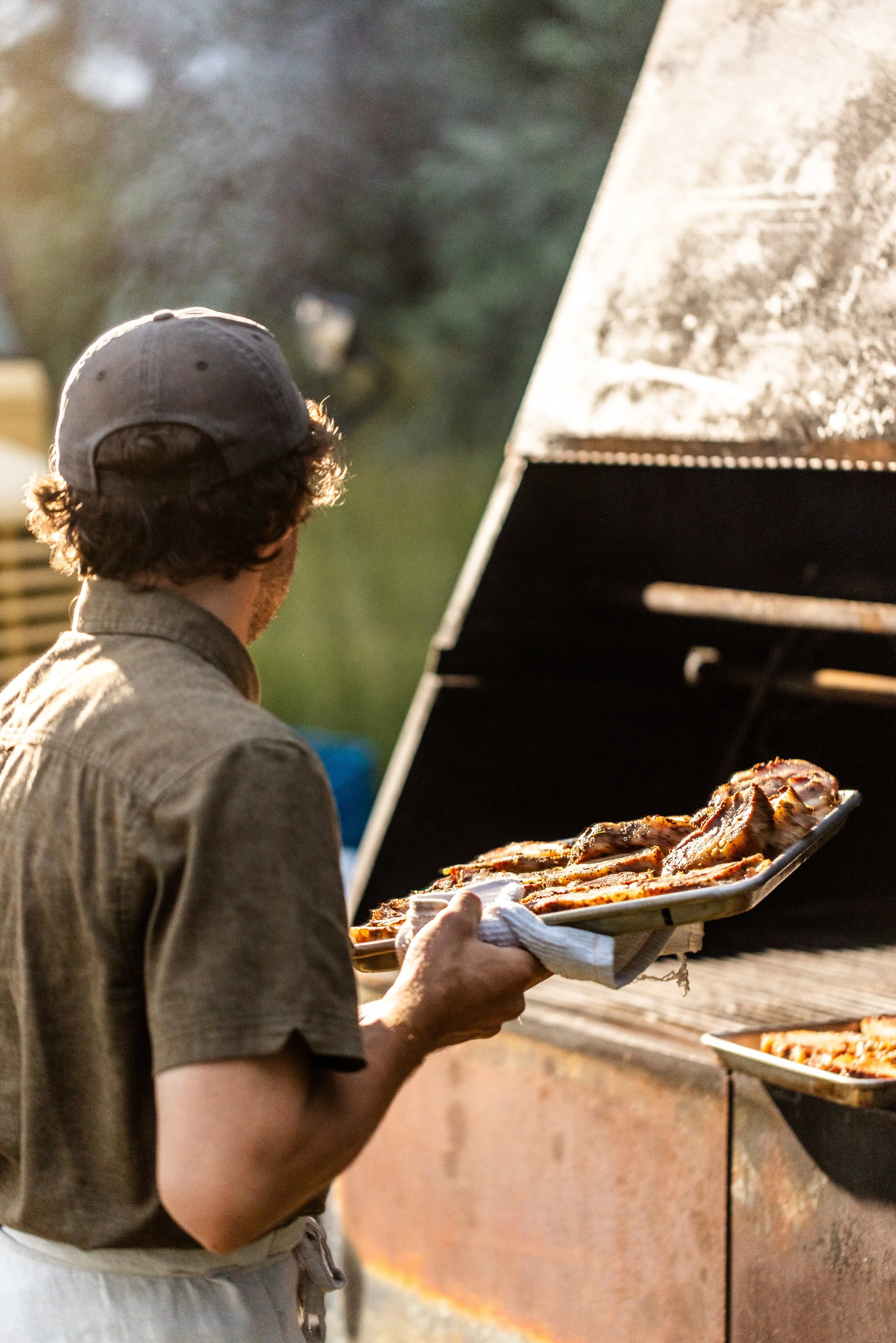 A man wearing a cap and apron is grilling food outside, holding a tray of cooked meat near an outdoor grill.