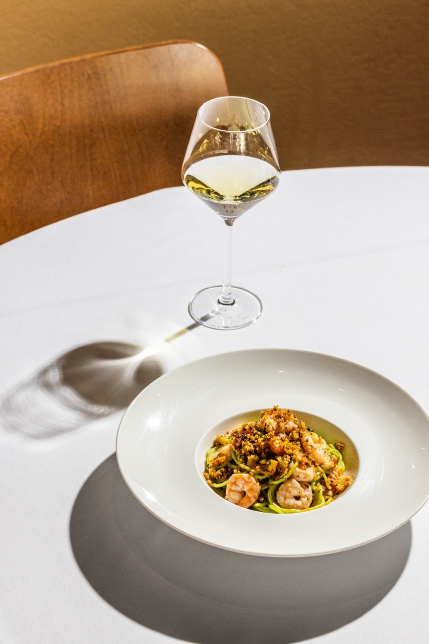 A glass of white wine on a white table with a plate of pasta, garnished with shrimp and crumbs, in a restaurant setting.