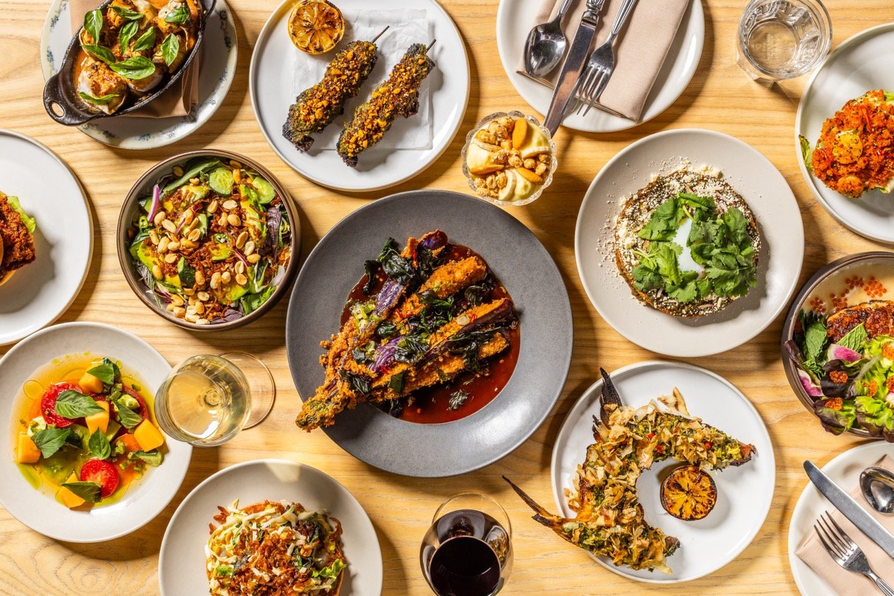 A variety of dishes on a wooden table including salads, fried fish with sauce, skewered appetizers, grilled vegetables, and glasses of white and red wine.