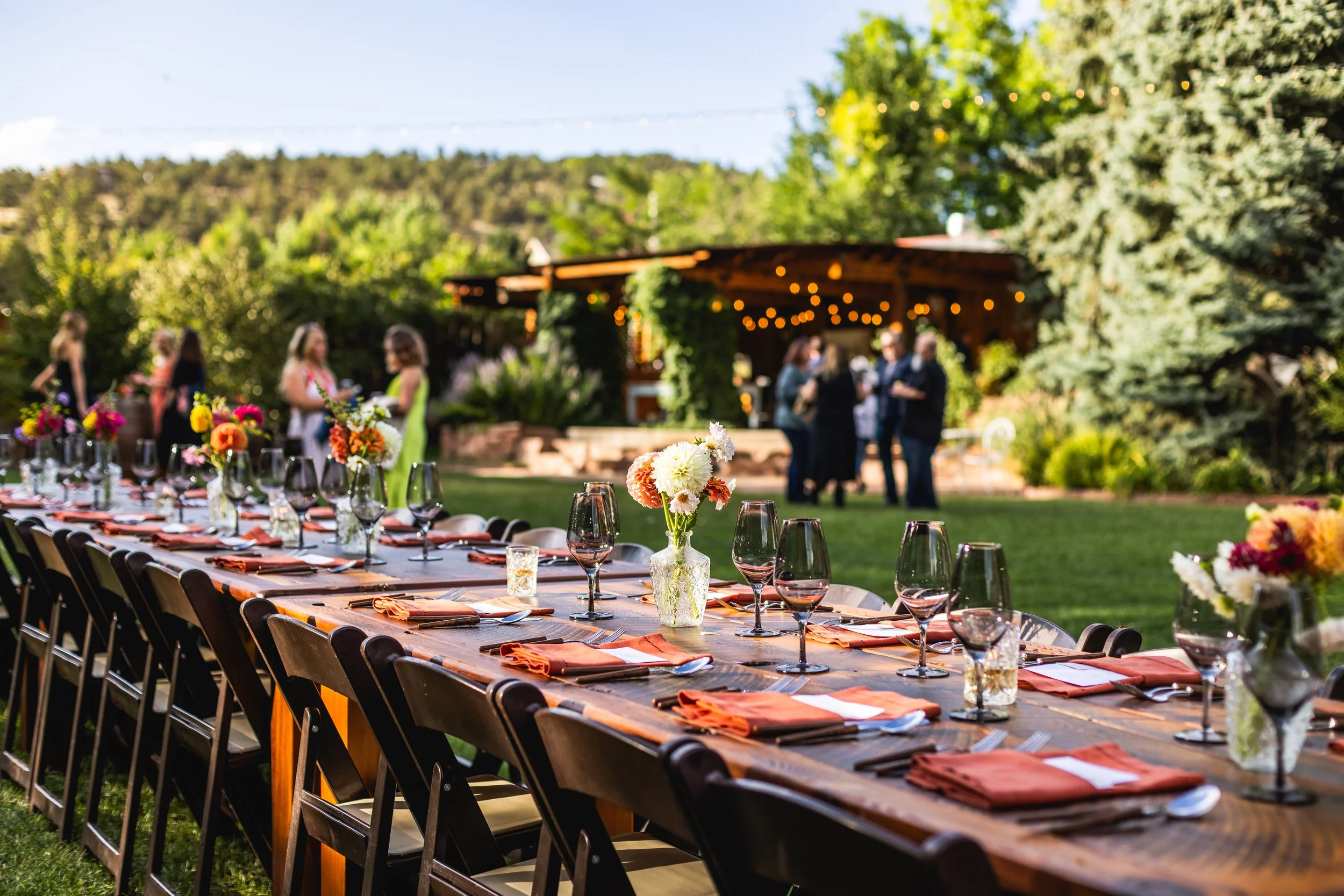 Long outdoor banquet table decorated with flowers, glassware, and napkins, set for a gathering, with people mingling in the background under string lights in a garden setting during daytime.