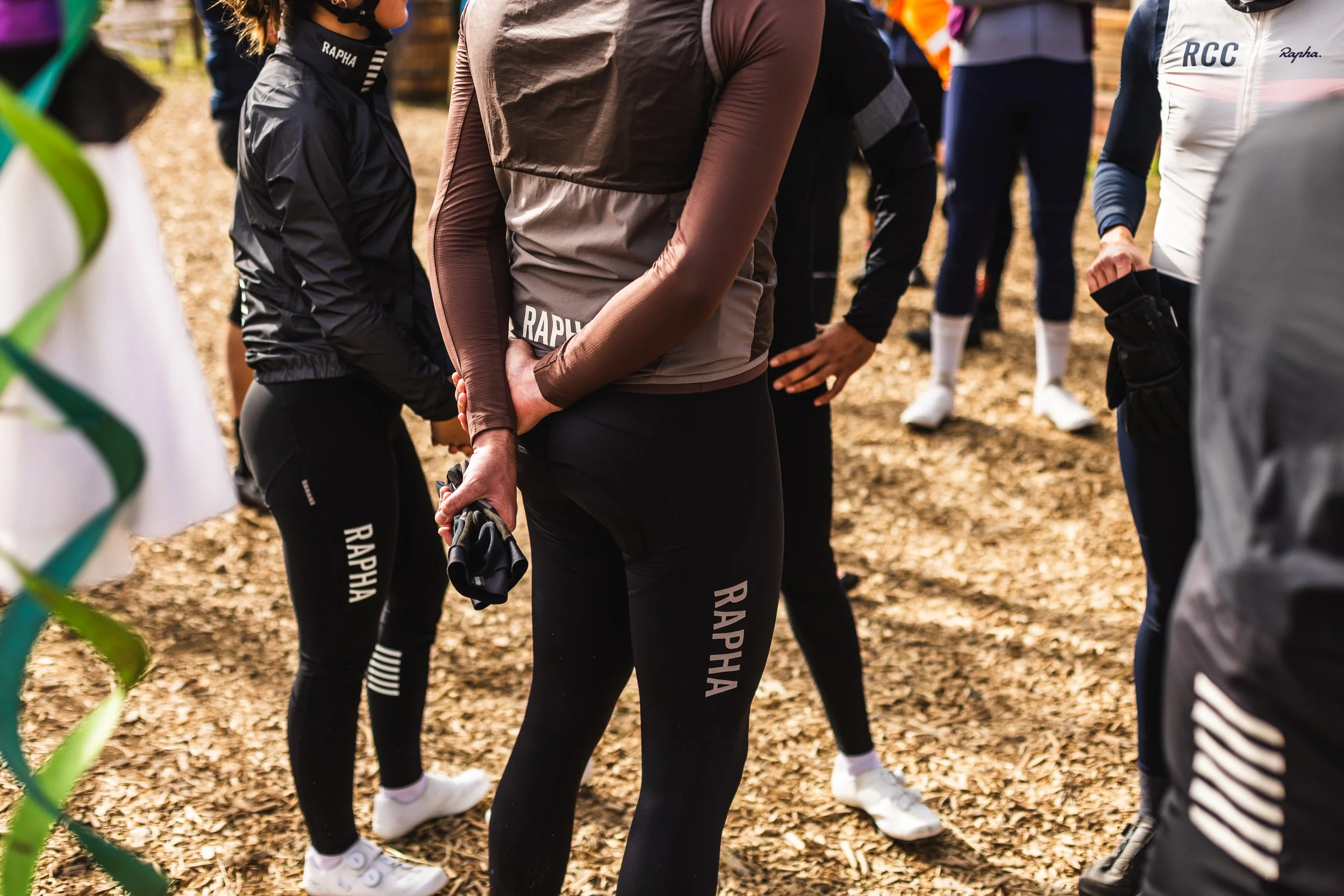 Group of cyclists in athletic gear gathered outdoors on a wooded trail, preparing for a race or training, with fallen leaves on the ground.