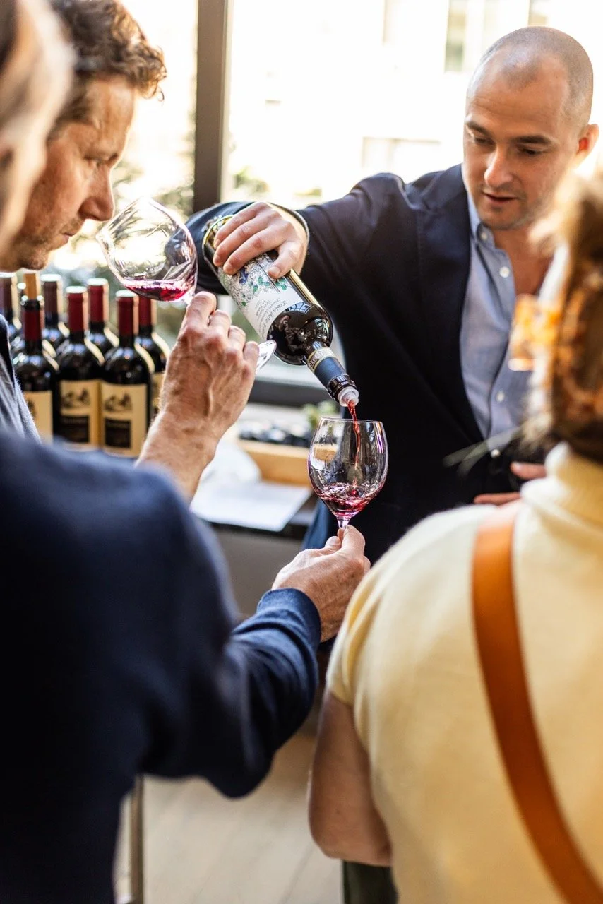 A sommelier pouring red wine into a glass while a man and woman observe.