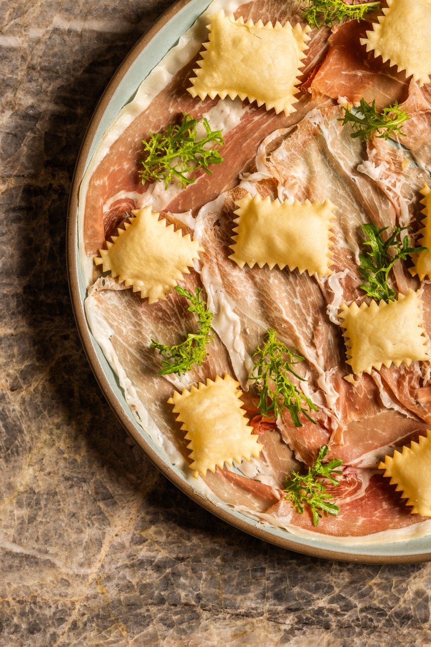 A close-up of a platter with raw slices of prosciutto, garnished with green parsley and topped with uncooked ravioli pasta on a dark marble surface.