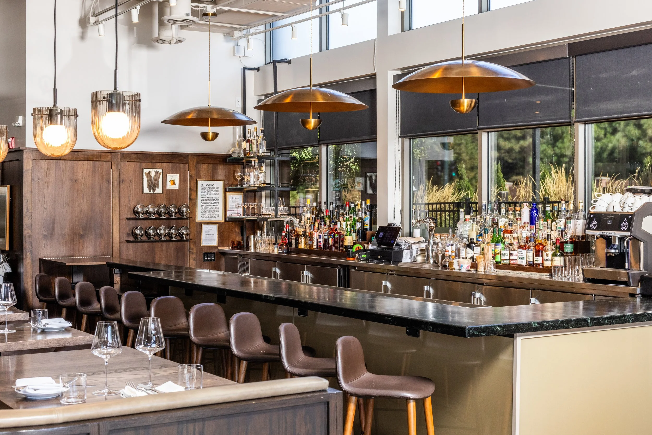 Modern bar with wooden and metal accents, pendant lights, high chairs, and a well-stocked liquor display near large windows showing outdoor greenery.