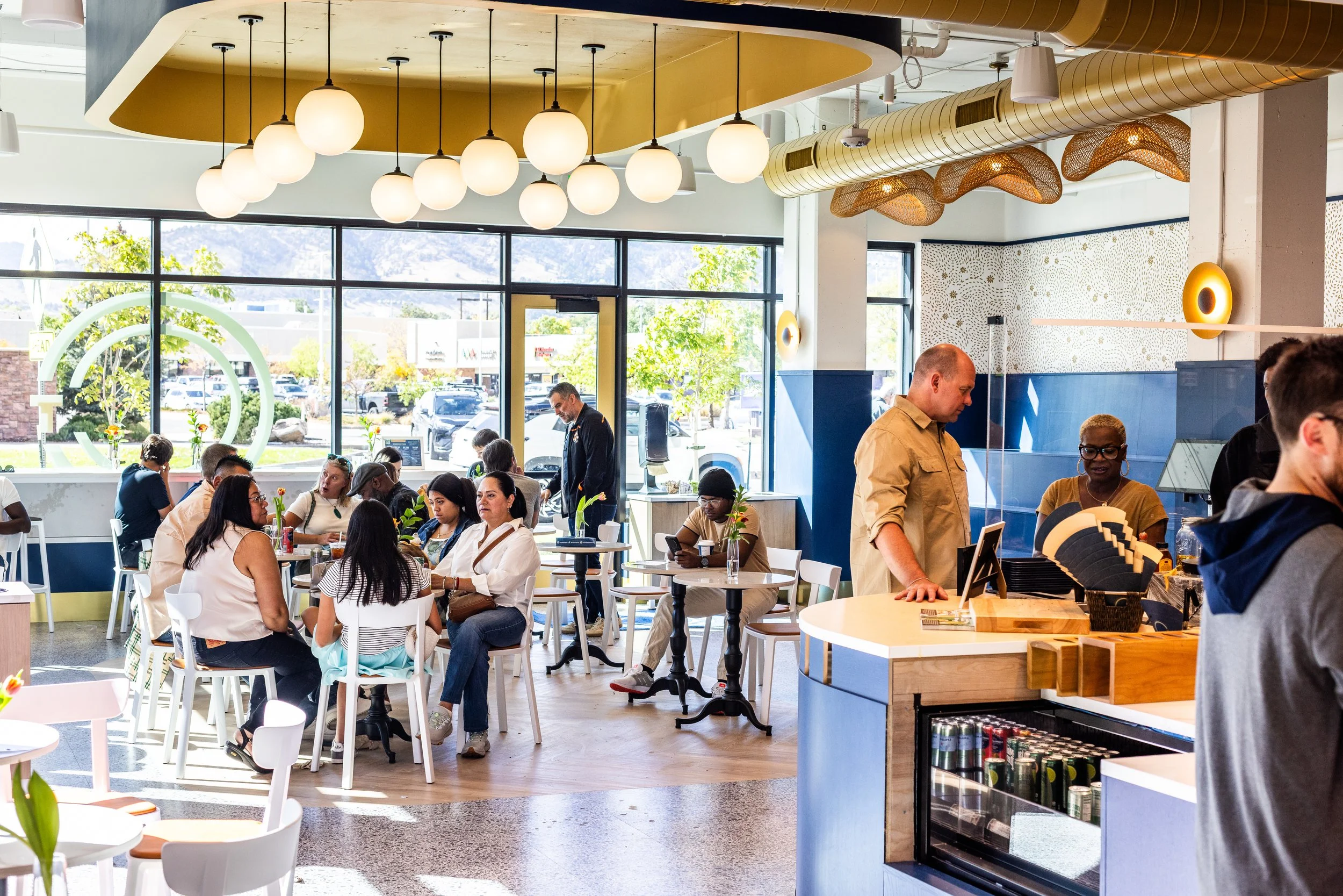 Inside a busy modern cafe with large windows, there are several groups of diverse customers sitting at tables, some using their phones. The counter area has a man and a woman staff member, and a young man in a gray hoodie is ordering. The decor featu