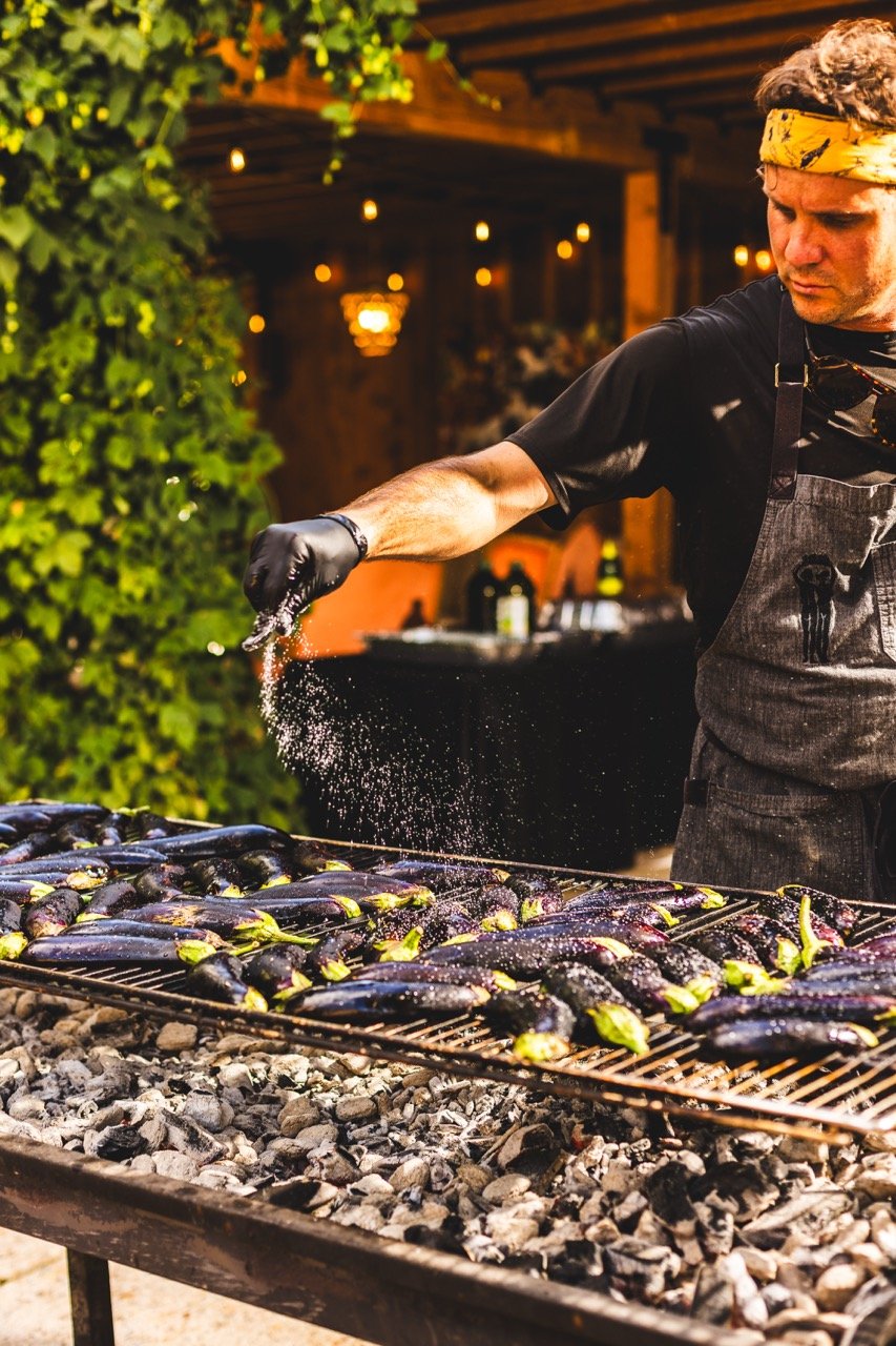A man wearing a yellow bandana and black gloves is seasoning eggplants on a grill outdoors during the evening, with blurred warm lighting and green foliage in the background.