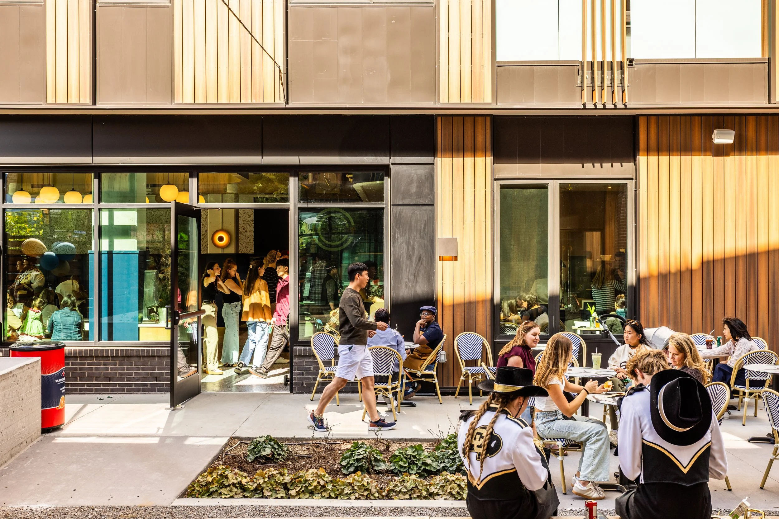 People dining outdoors at tables in front of a modern building with large glass windows and wooden paneling, some people standing in line inside the restaurant.