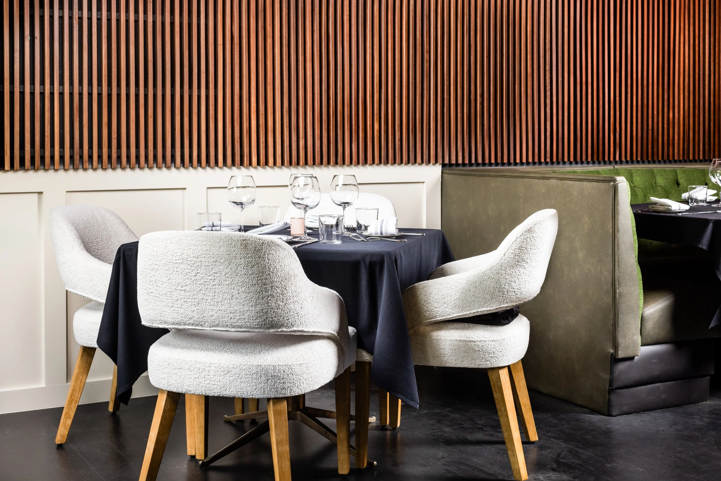 A restaurant table set for dining with four white cushioned chairs, a black tablecloth, wine glasses, water glasses, and neatly folded napkins, against a backdrop of a wooden slat wall and green upholstered banquette seating.