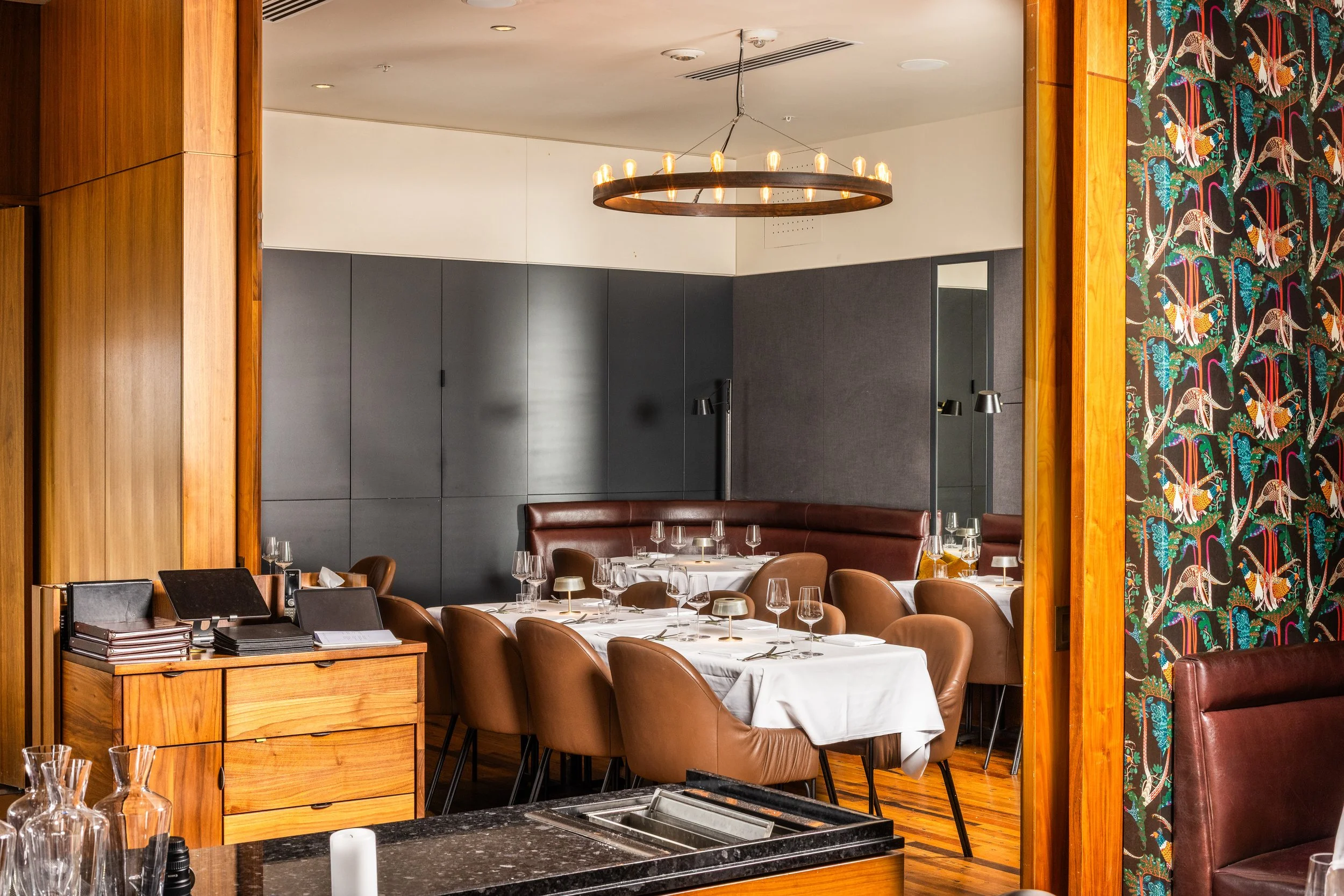 A dining room with a long table set for a meal, featuring white tablecloths, wine glasses, and cutlery. The room has brown leather chairs, a wooden sideboard with books and a black object, and decorative wallpaper with birds. A circular chandelier ha