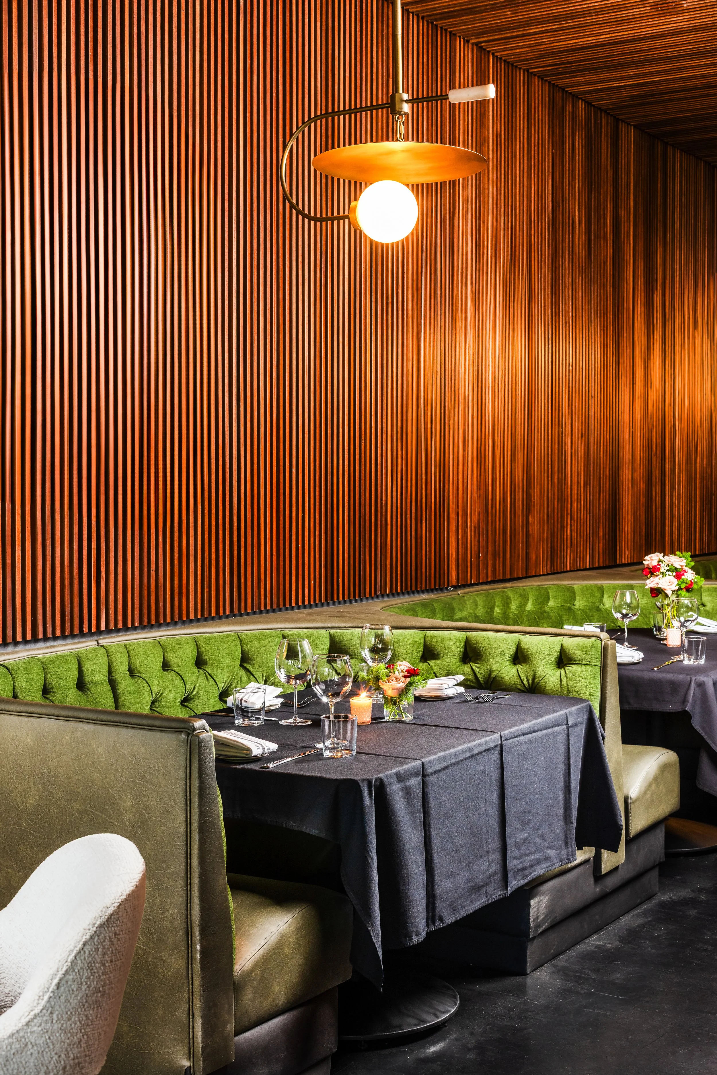 Restaurant dining area with black tablecloths, green upholstered banquettes, floral centerpieces, wine glasses, candles, and a wooden slat wall with warm lighting.