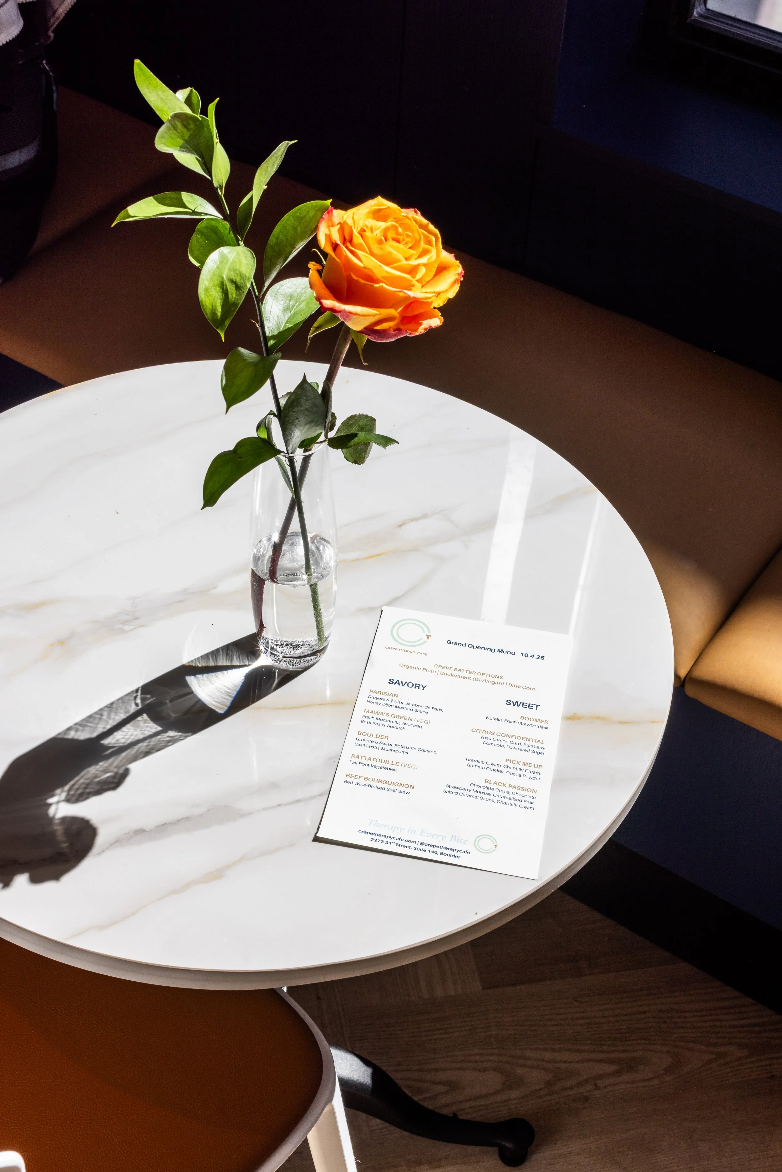 A single orange and yellow rose in a glass vase on a white marble table with a menu and shadow.