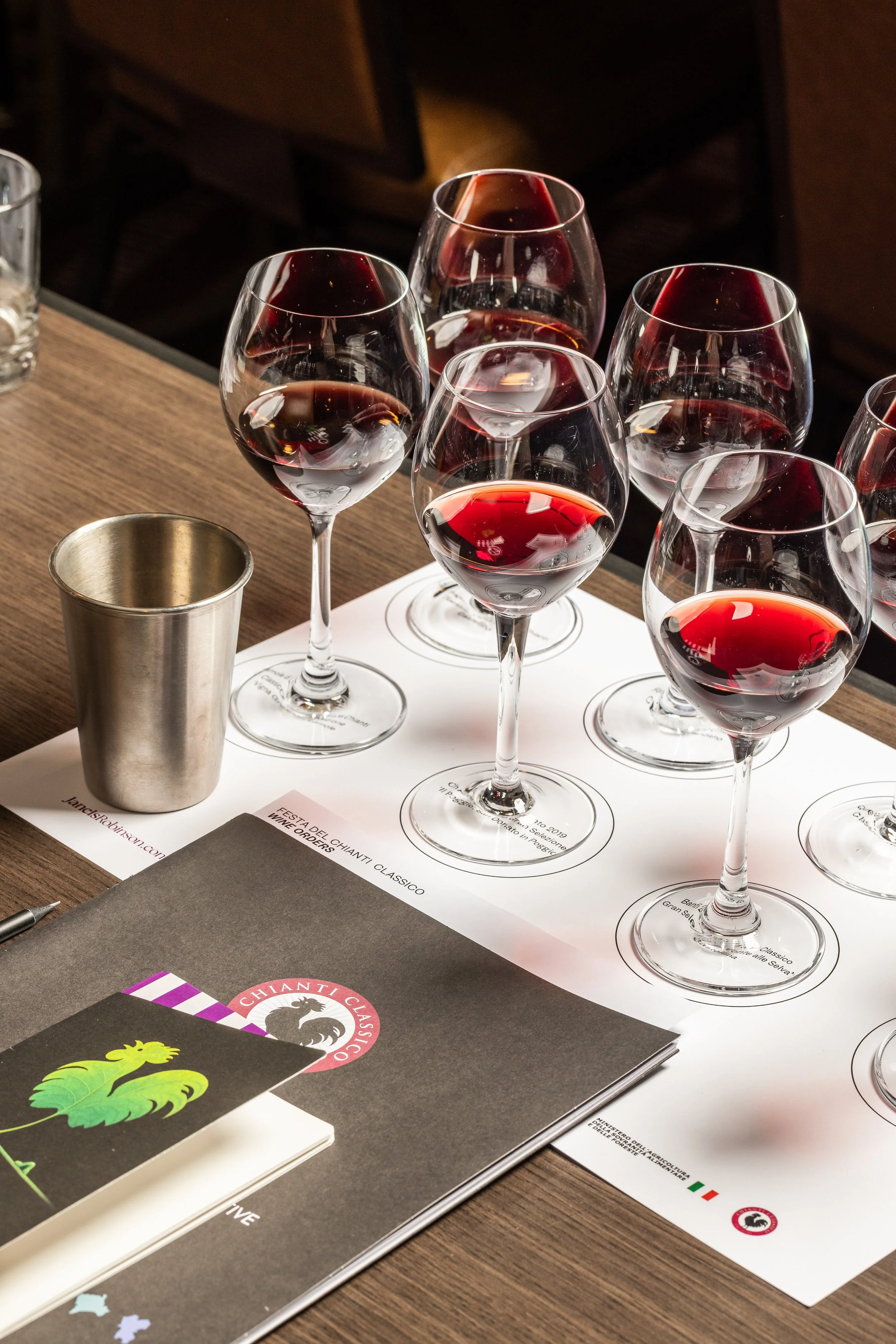 Several glasses of red wine on a tasting sheet with a booklet and metal cup on a wooden table.