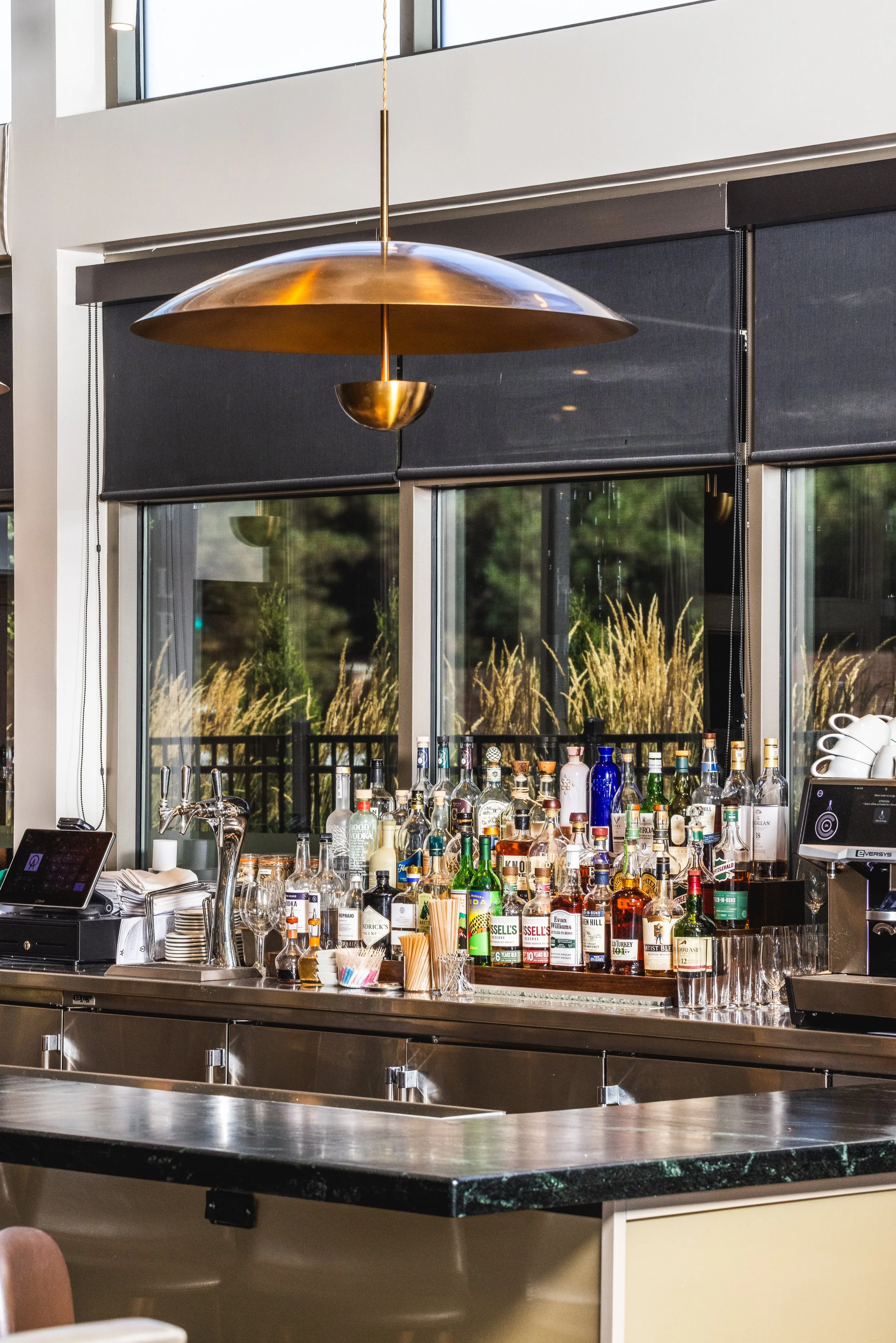 View of a bar with various bottles of alcohol, glasses, and bar tools, with a window showing an outdoor scene with trees and grasses.
