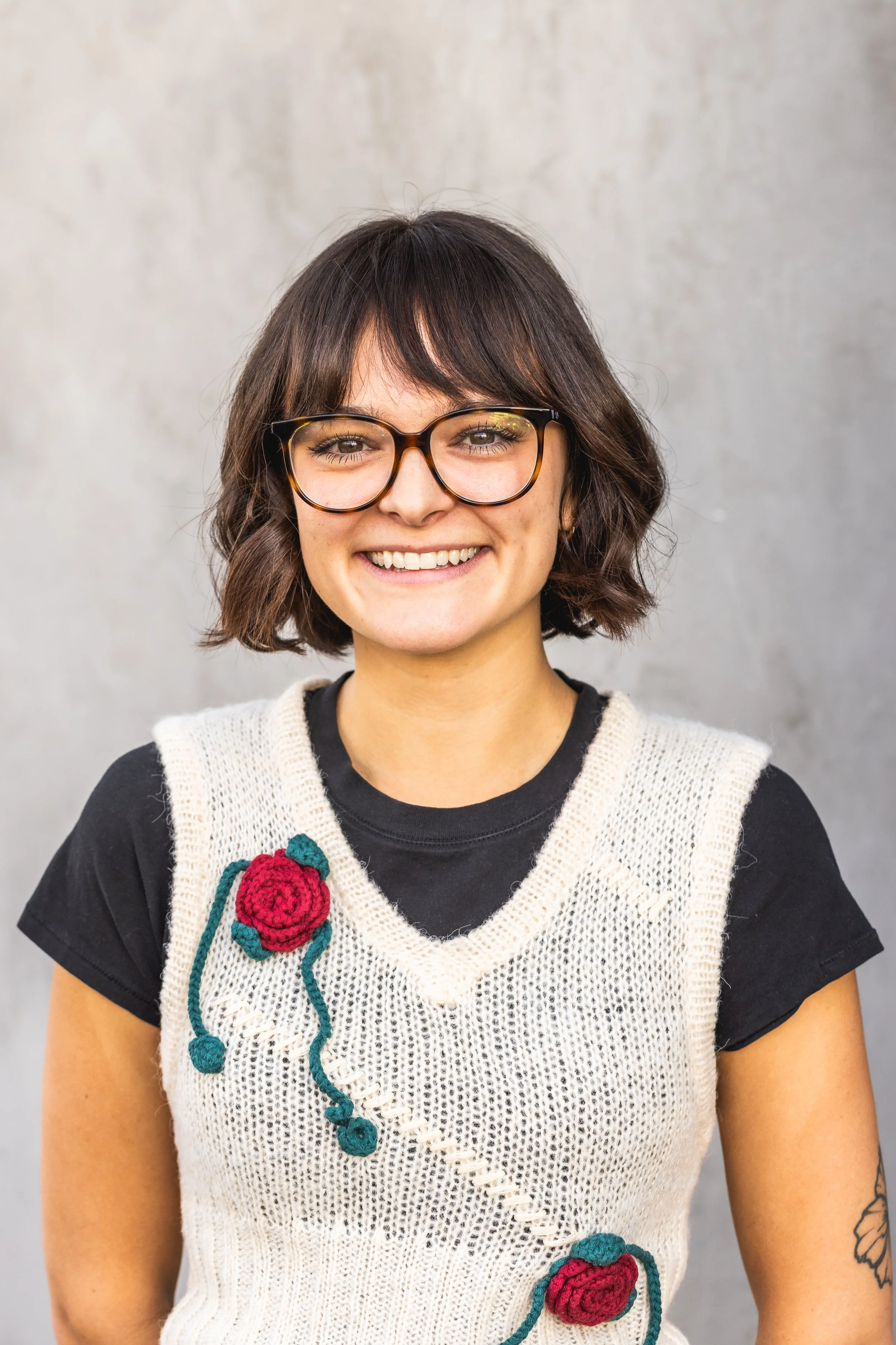 A woman with glasses and short brown hair, smiling, wearing a knitted cream vest with red flower embroidery over a black t-shirt, standing against a gray background.