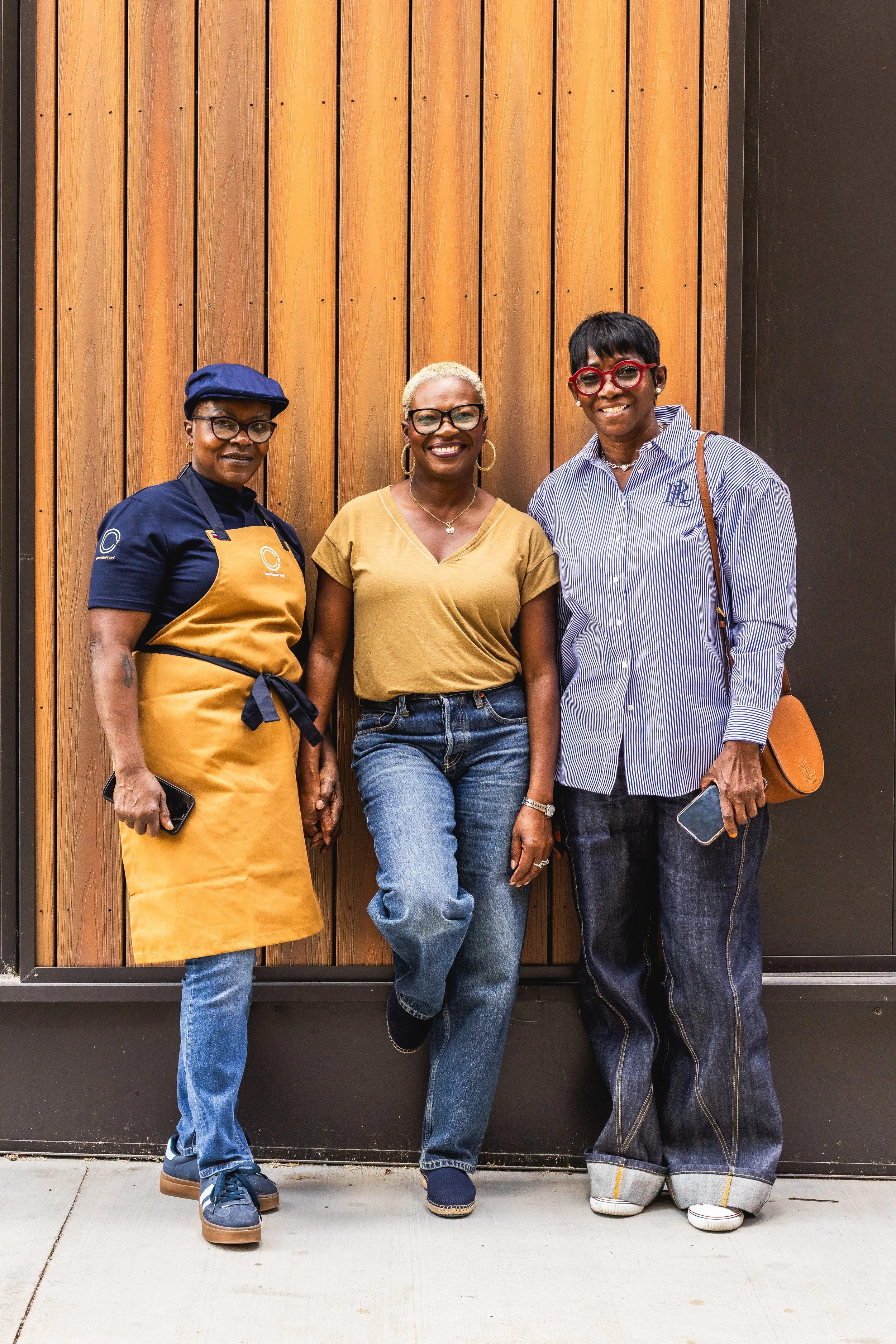 Three women standing together against a wooden wall. The woman on the left is wearing a navy shirt, mustard apron, blue cap, and jeans. The woman in the middle has short blonde hair, wearing a mustard T-shirt and jeans. The woman on the right has sho