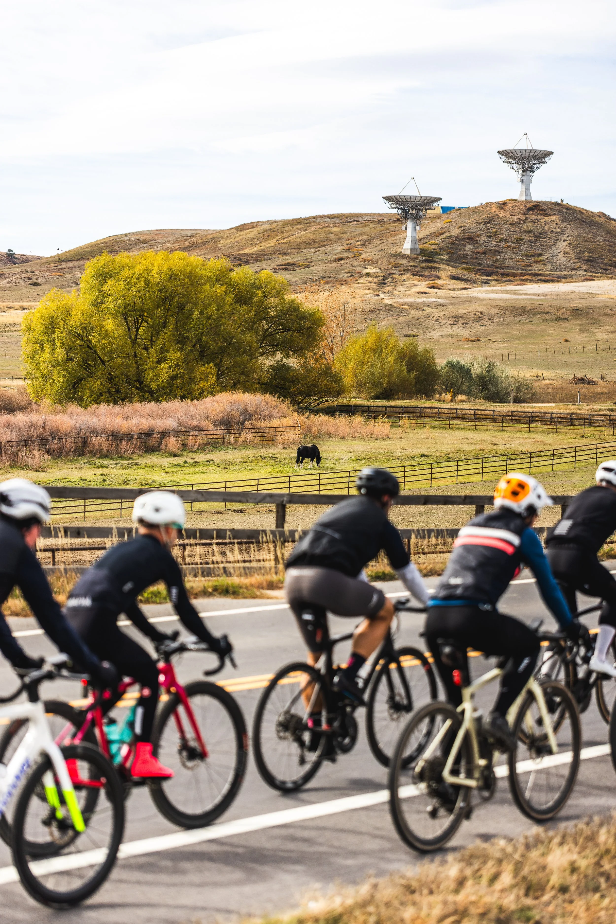 Cyclists riding on a road with hills, trees, and radio telescopes in the background.