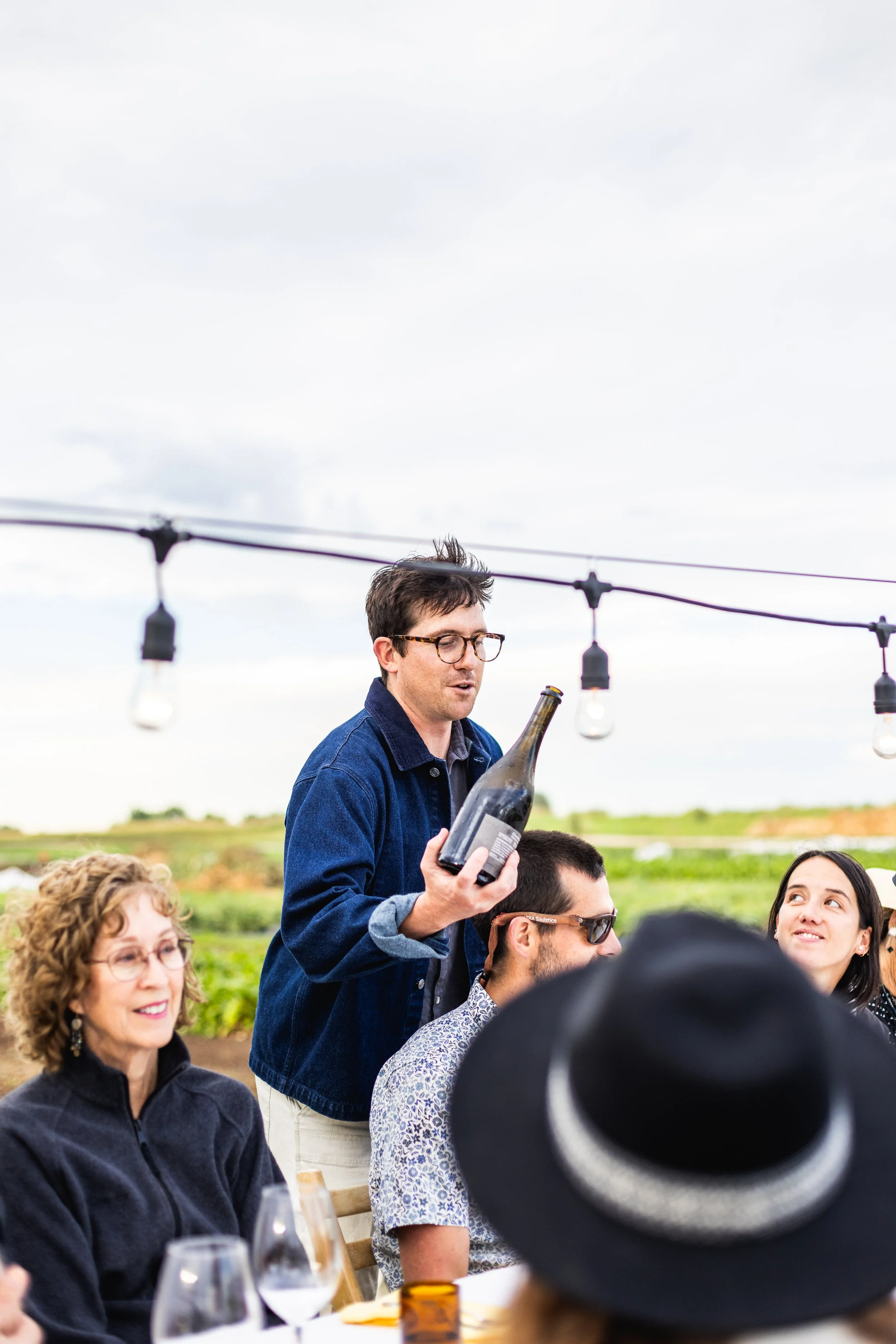 A man with glasses standing outdoors at a gathering, holding a bottle, while people seated around a table listen. The background shows a cloudy sky and an open field with string lights hanging overhead.