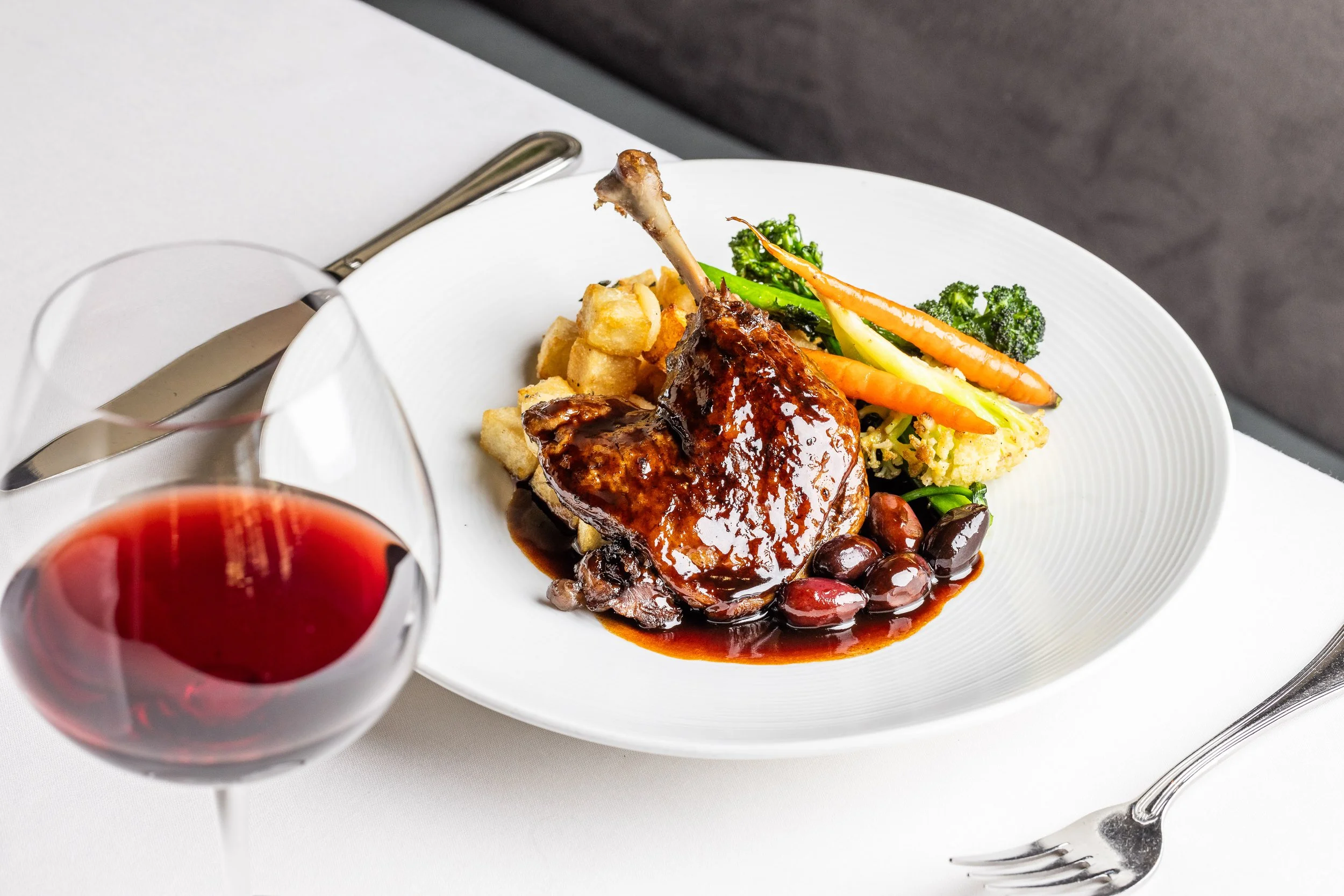 Plate with glazed roasted rack of lamb, roasted potatoes, steamed broccoli, carrots, cauliflower, and roasted olives, with a glass of red wine beside it on a white tablecloth.