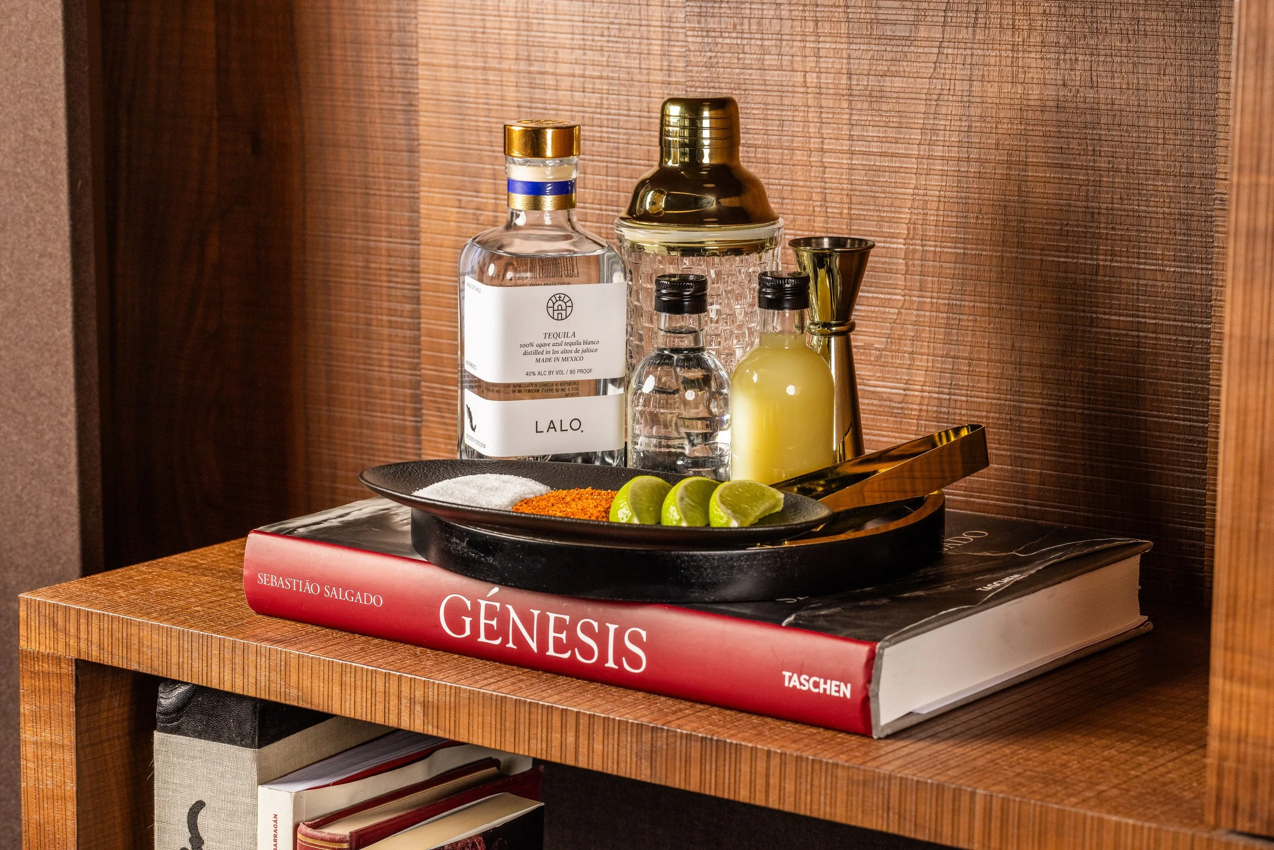 A wooden side table with a large book titled 'GÉNESIS' and a black decorative tray holding lime wedges, salt, and chili powder, with various liquor bottles including tequila and aquavit, a cocktail shaker, and a small measuring jigger.