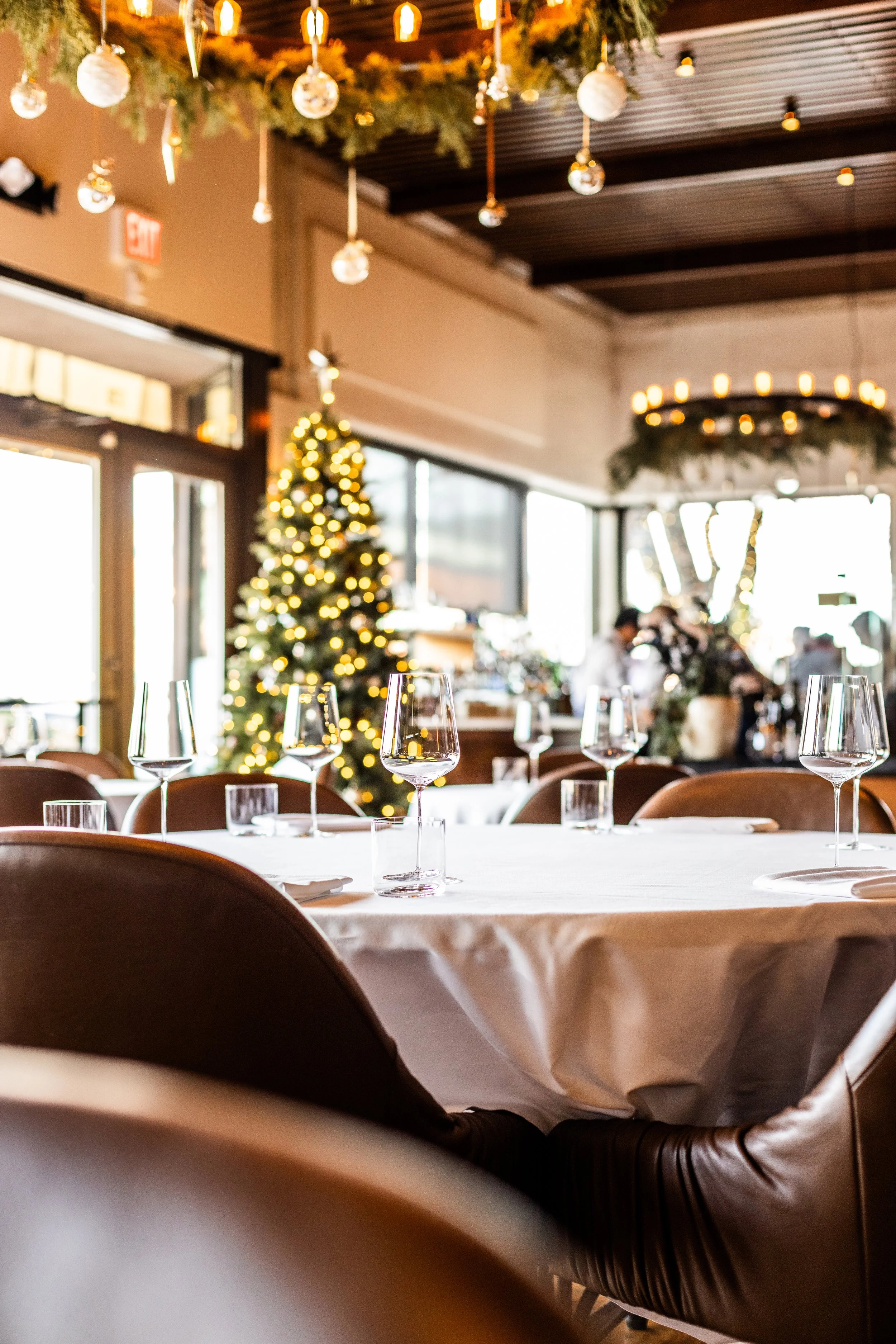 A decorated restaurant interior with a Christmas tree, hanging ornaments, and a chandelier, with empty table settings in the foreground.