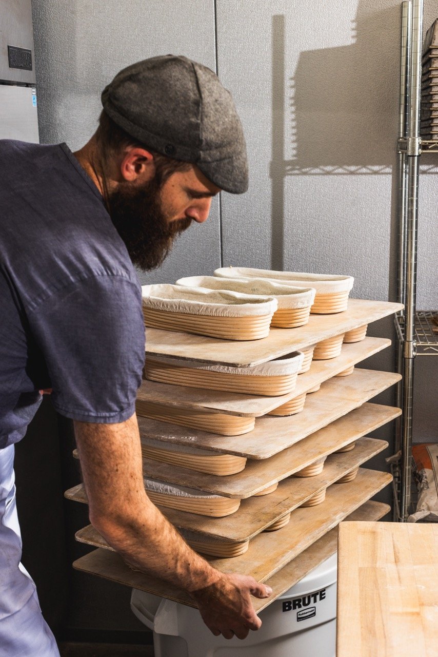 A man with a gray cap and a beard places wooden trays with fabric-lined molds on top of a white container in a workshop setting.