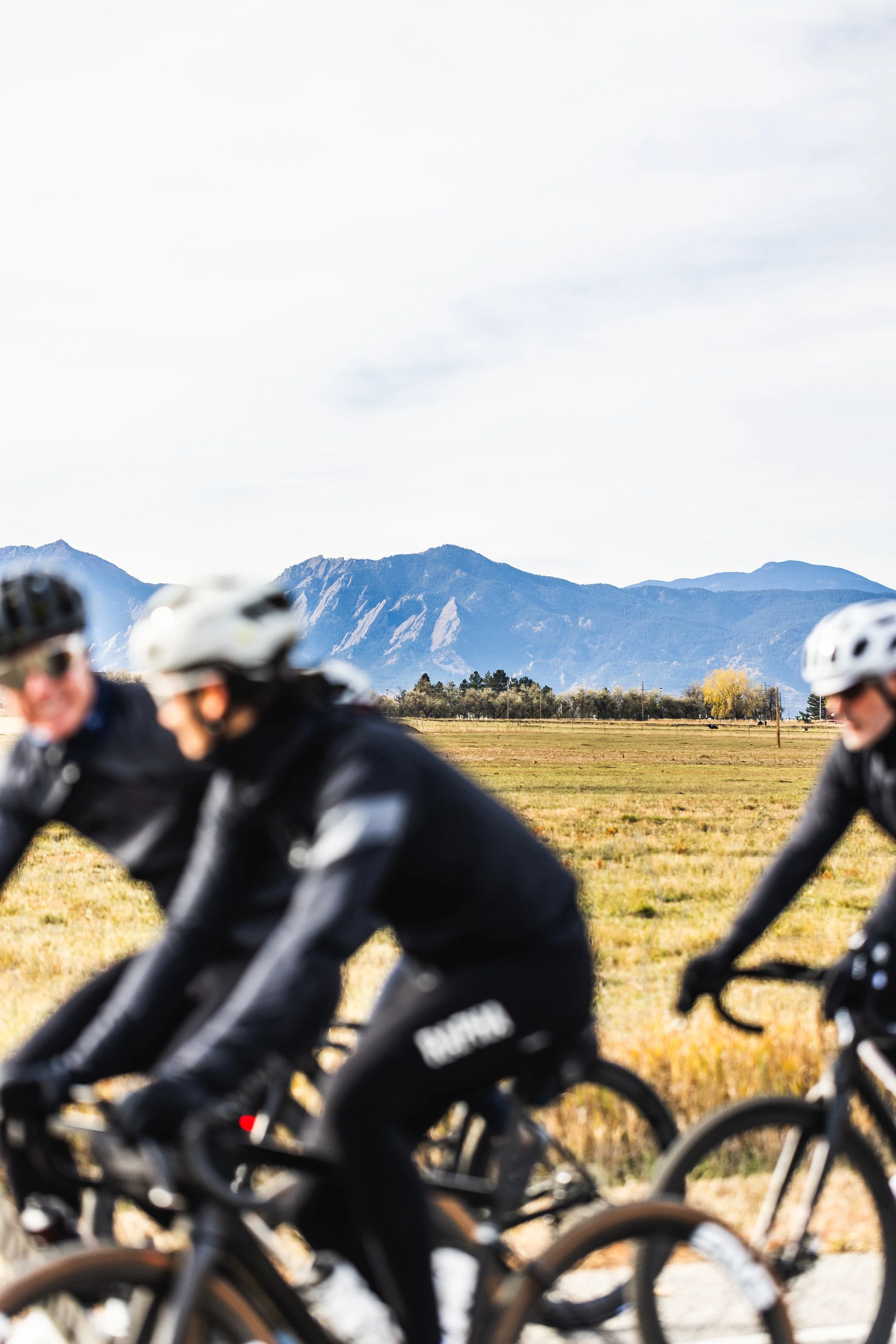 A group of cyclists riding mountain bikes on a trail in a rural area with mountains in the background, wearing helmets and black cycling gear.