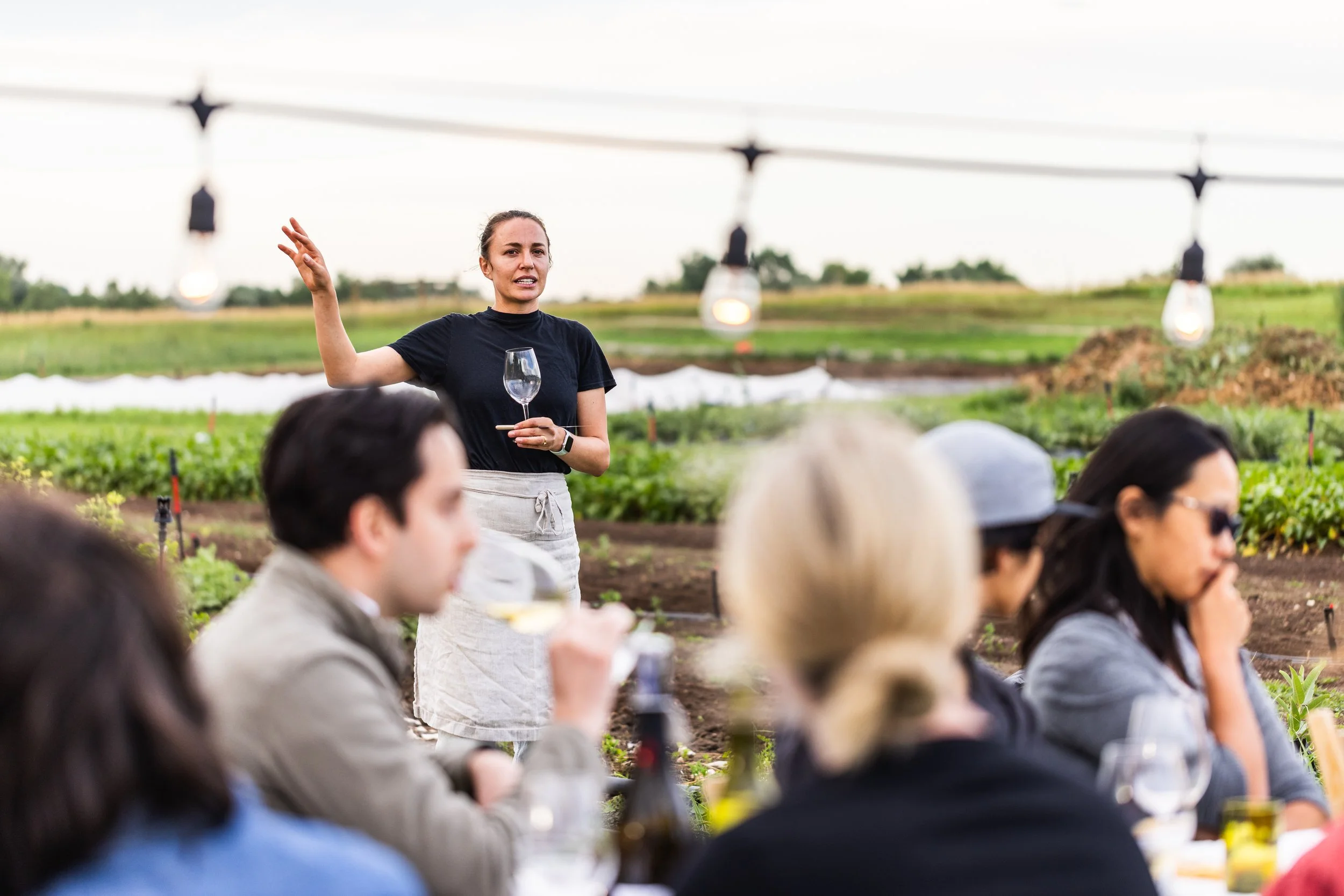 A woman giving a presentation or speech to a group of people outdoors in a farm or garden setting, with string lights hanging above.