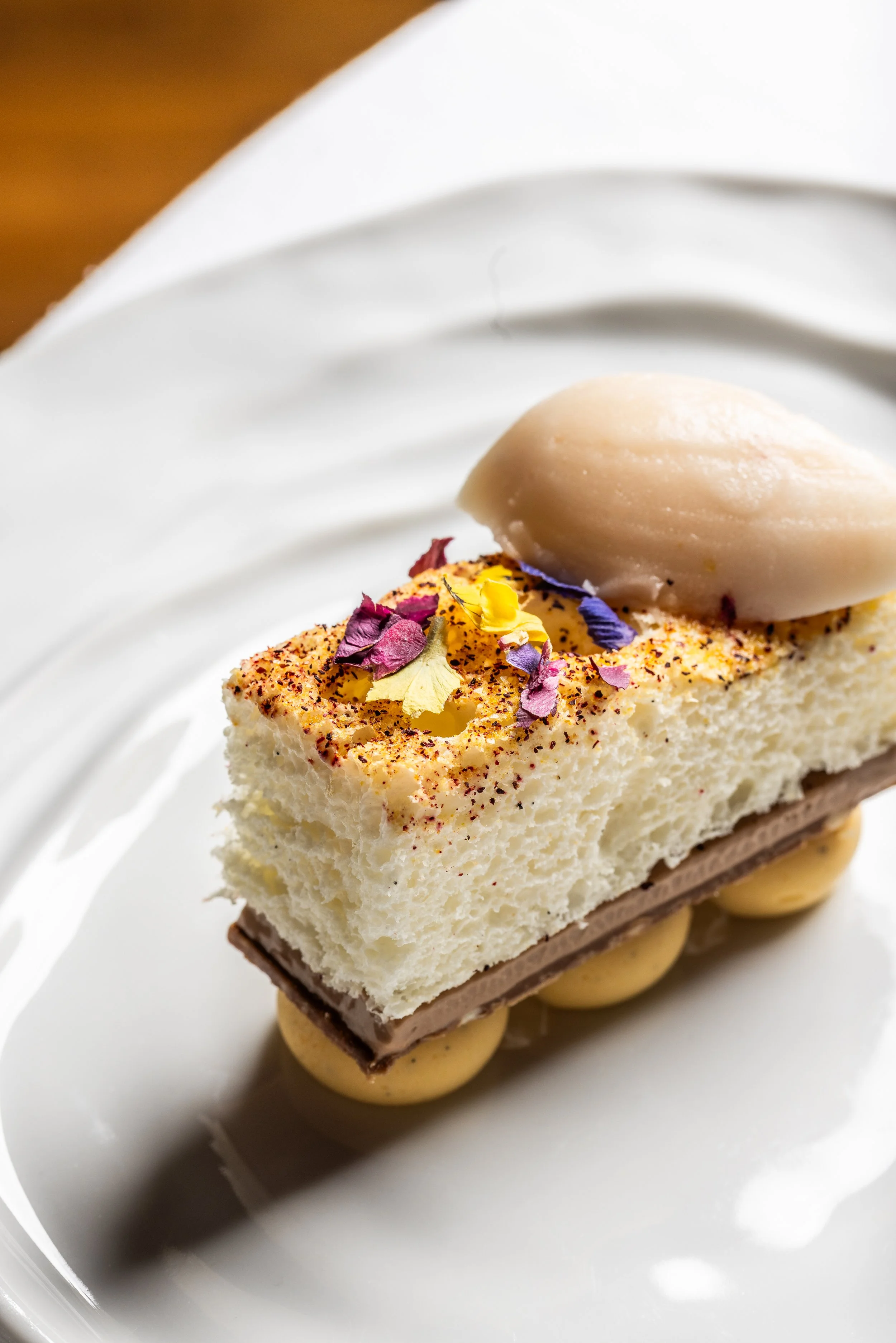 A slice of layered dessert with whipped cream, edible flower petals, and a scoop of ice cream or sorbet on top, served on a white plate.