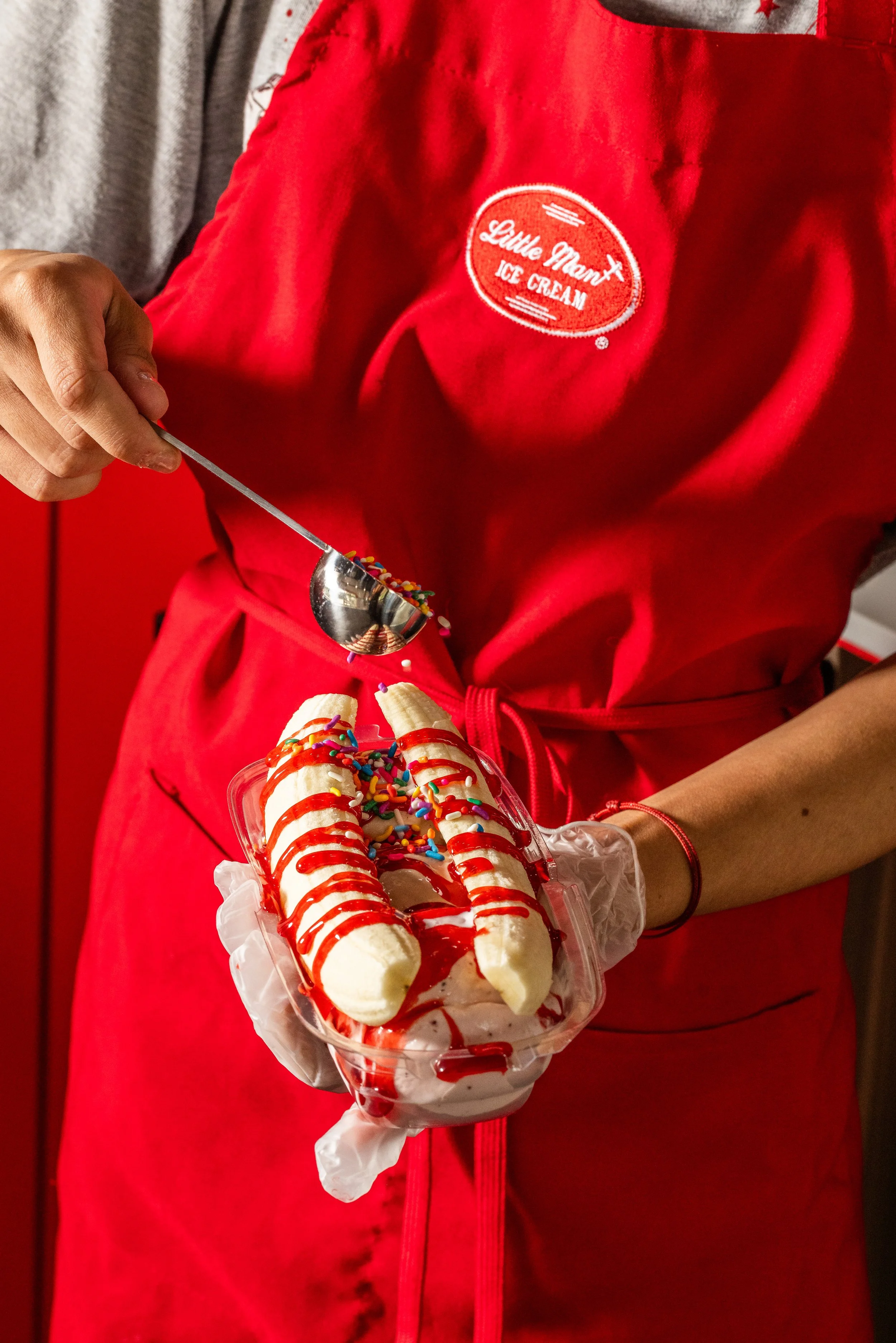 Person wearing a red apron holding a cup of banana split topped with whipped cream, rainbow sprinkles, and red syrup.