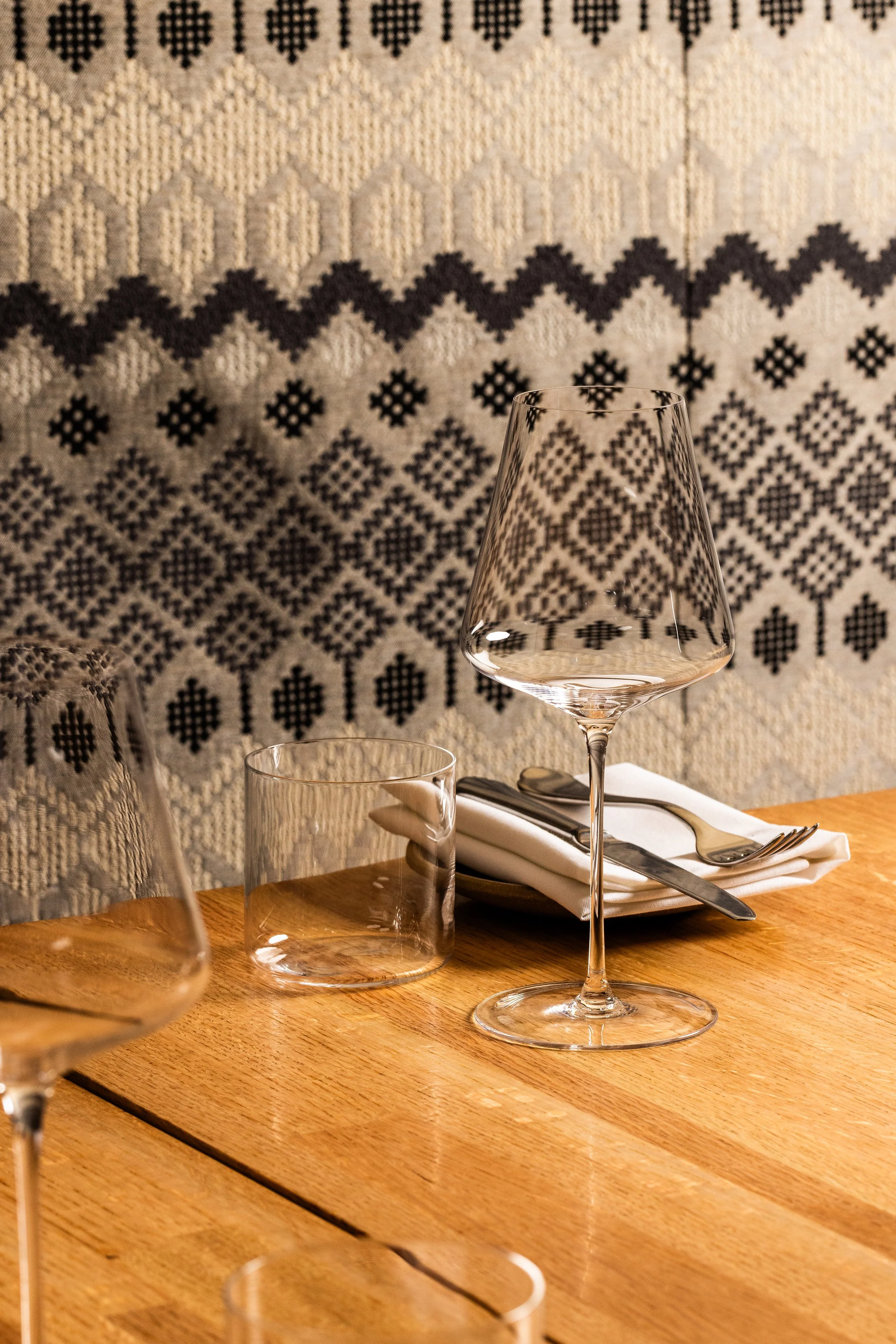 Empty wine glass, water glass, and set of silverware on a folded napkin on a wooden table in front of a patterned textile background.