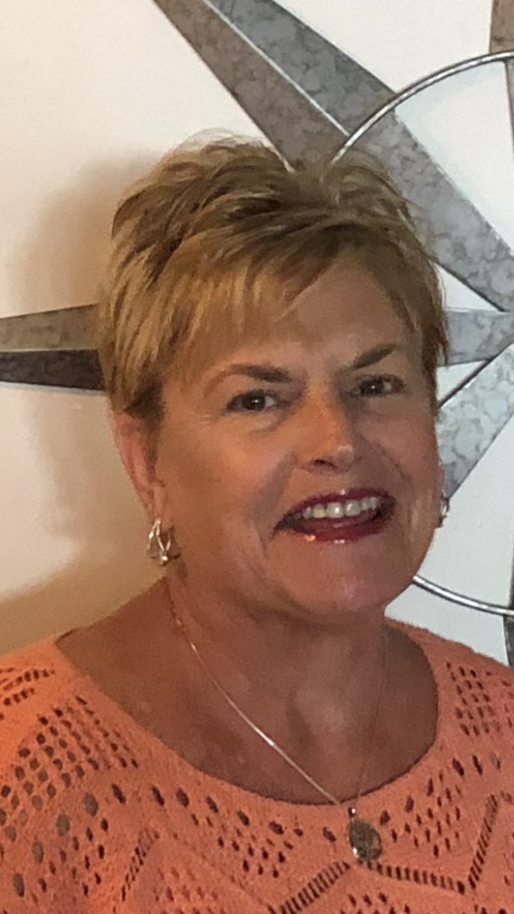 A smiling middle-aged woman with short blonde hair, wearing a peach-colored crochet top, earrings, and a necklace with a pendant. She is standing in front of a wall with a modern decorative piece and a marble surface.