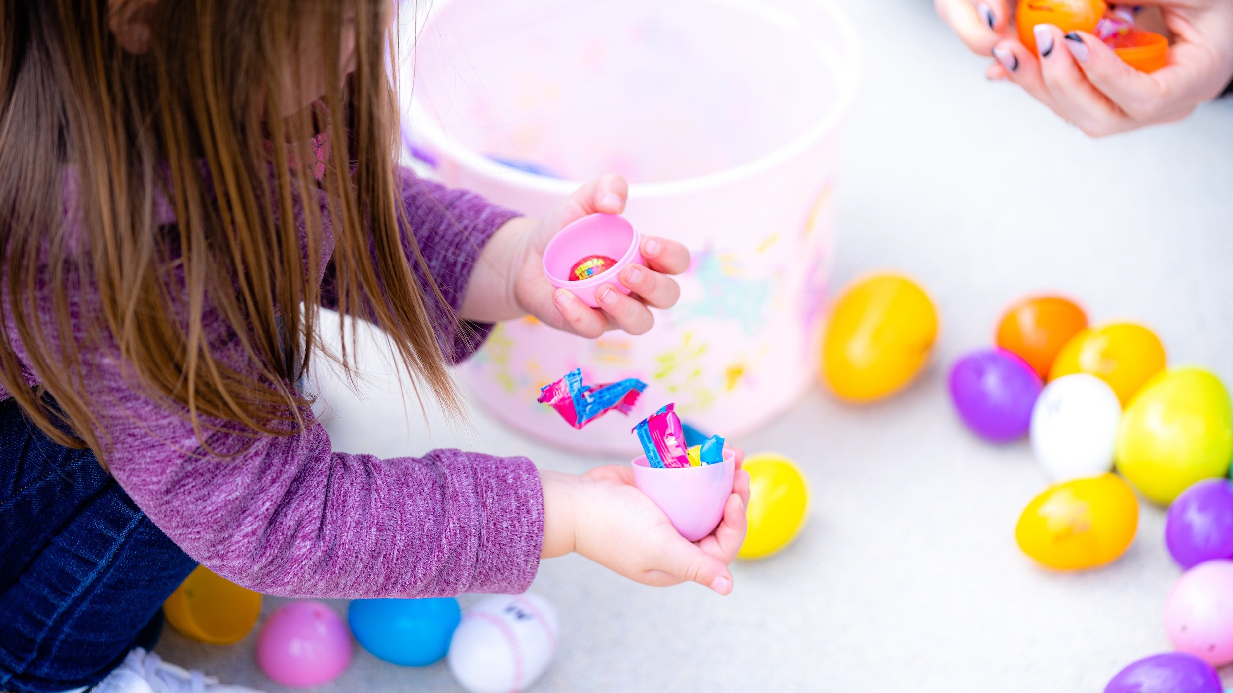 Child with long hair and purple sweater opening a pink plastic egg filled with colorful candy, surrounded by pastel-colored plastic eggs.