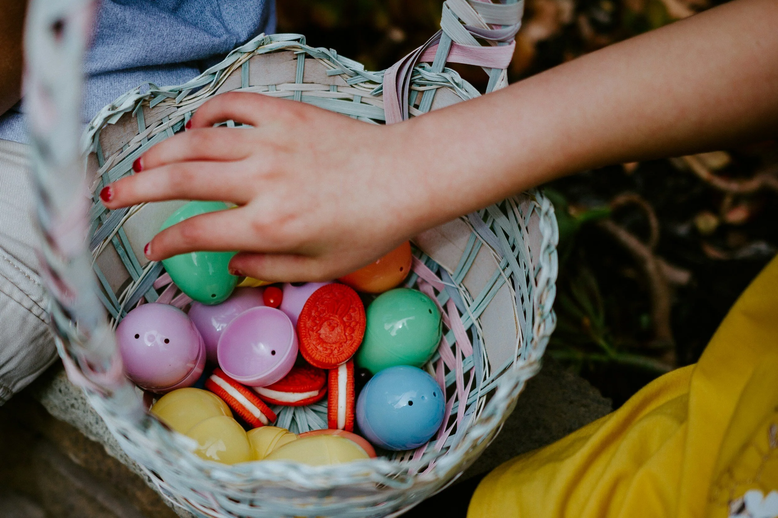 A child's hand reaching into a basket filled with colorful plastic Easter eggs and candies.