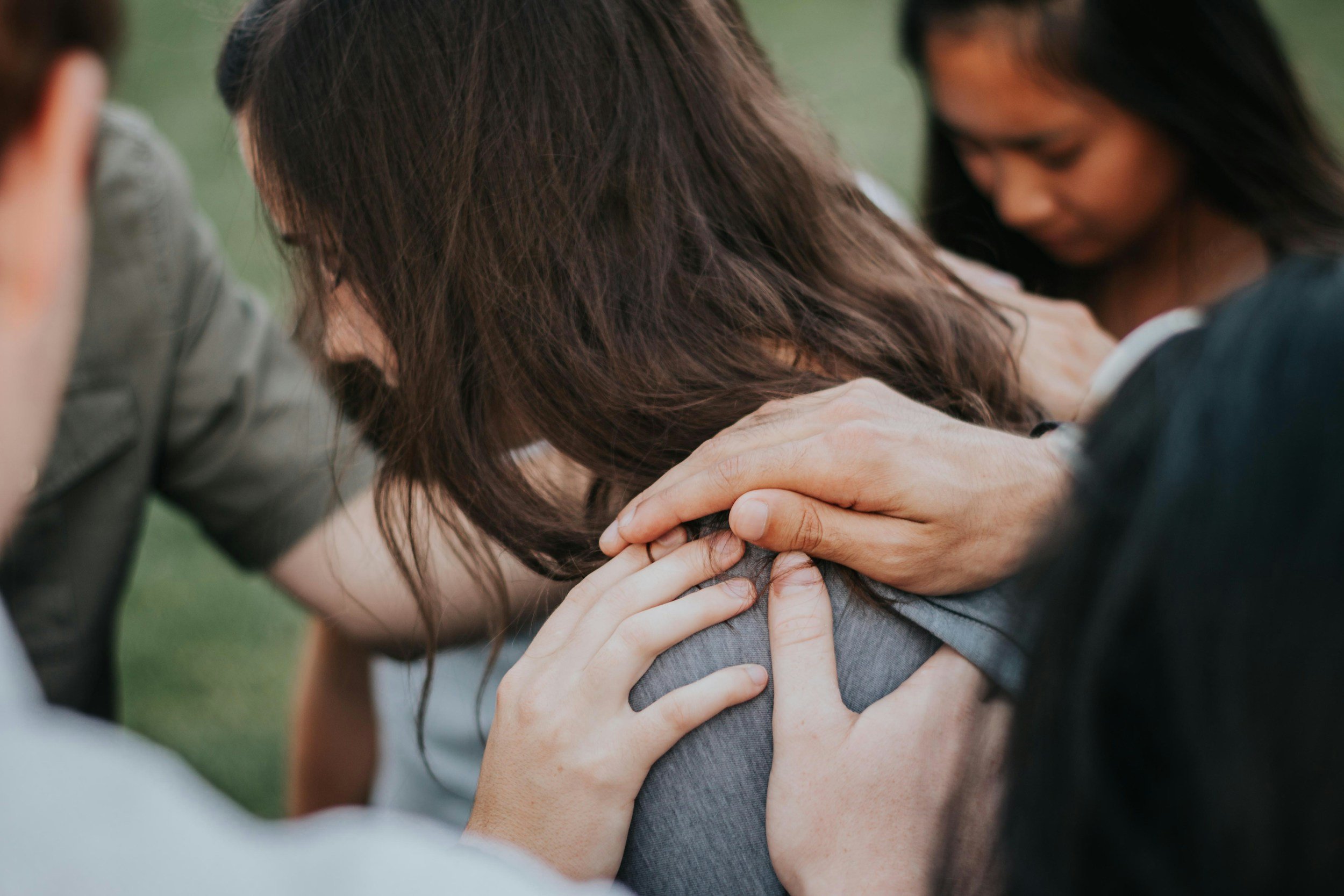 A group of people in a close, kneeling circle, with hands placed on each other's shoulders in a gesture of support and prayer.