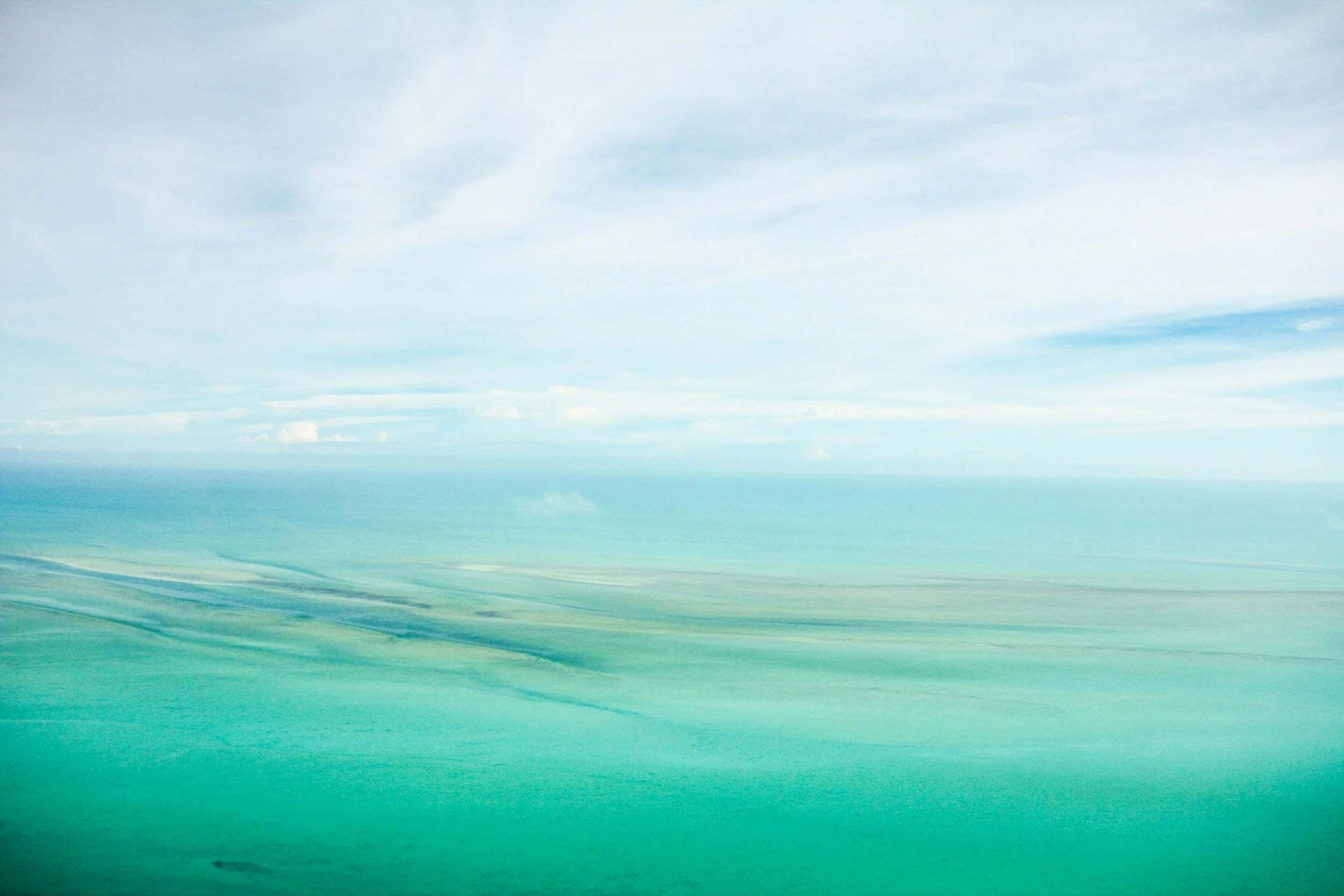 Aerial view of the ocean with varying shades of turquoise and blue, and partly cloudy sky above.
