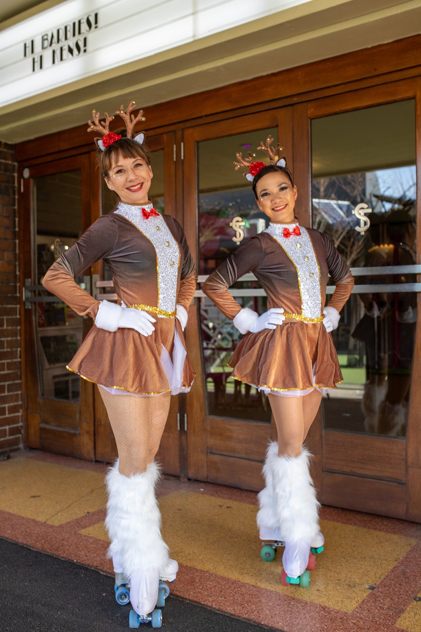 Two people in reindeer costumes with red bows, white gloves, and roller skates, standing in front of a wooden door. Sign reads "Hi Barbies! Hi Kens!"