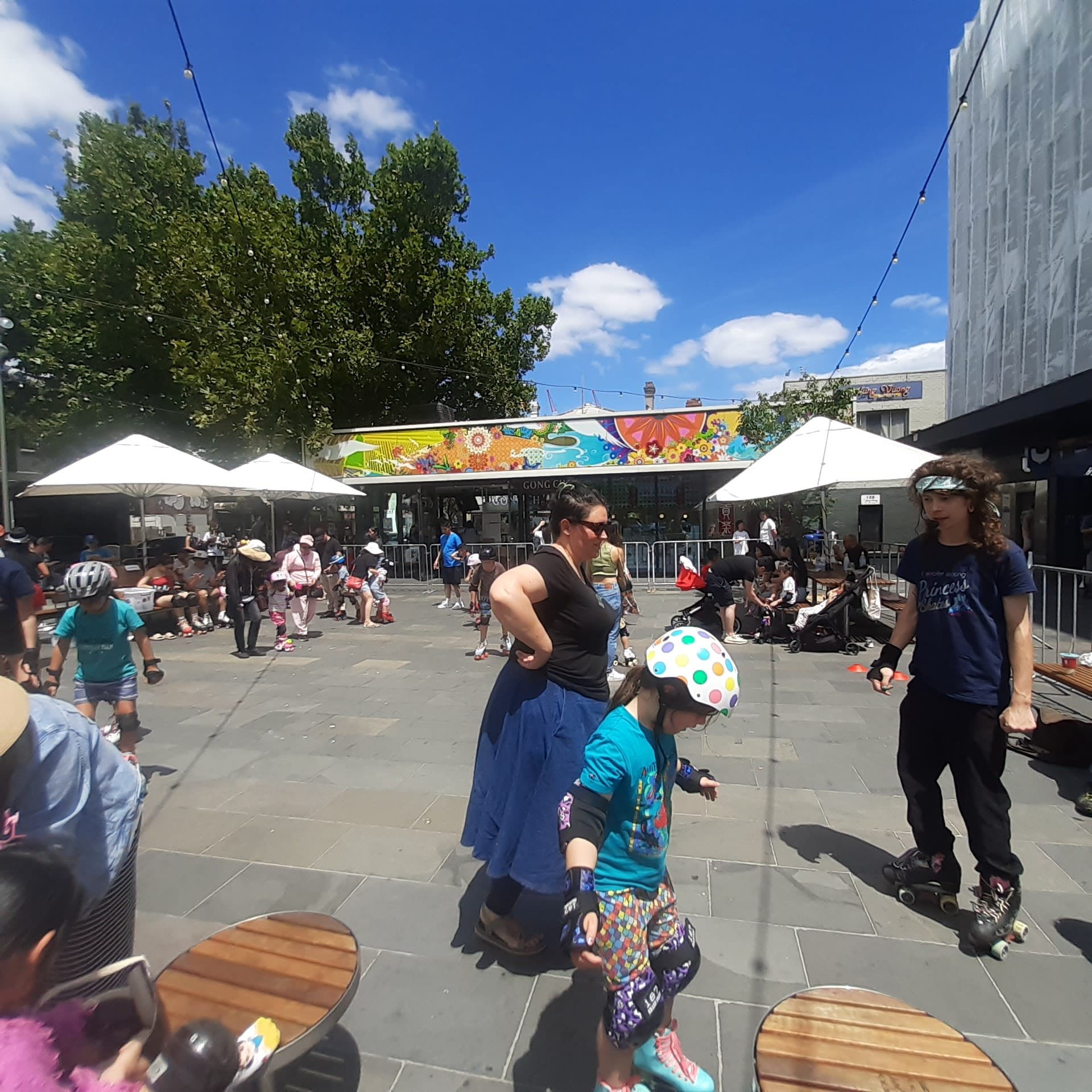 People roller skating in an outdoor plaza with vibrant mural and trees, sunny day, social gathering