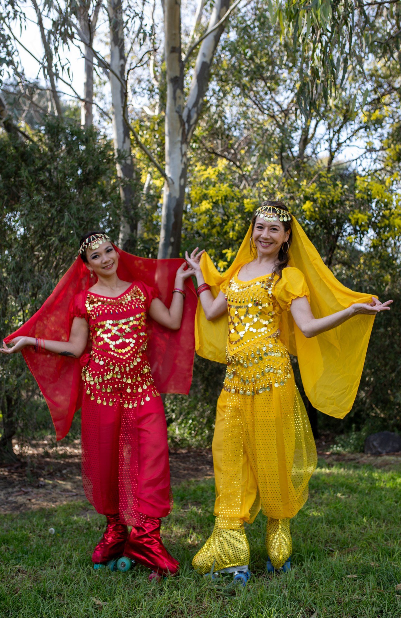 Two women in vibrant belly dance costumes, one in red and the other in yellow, standing outdoors in a wooded area.