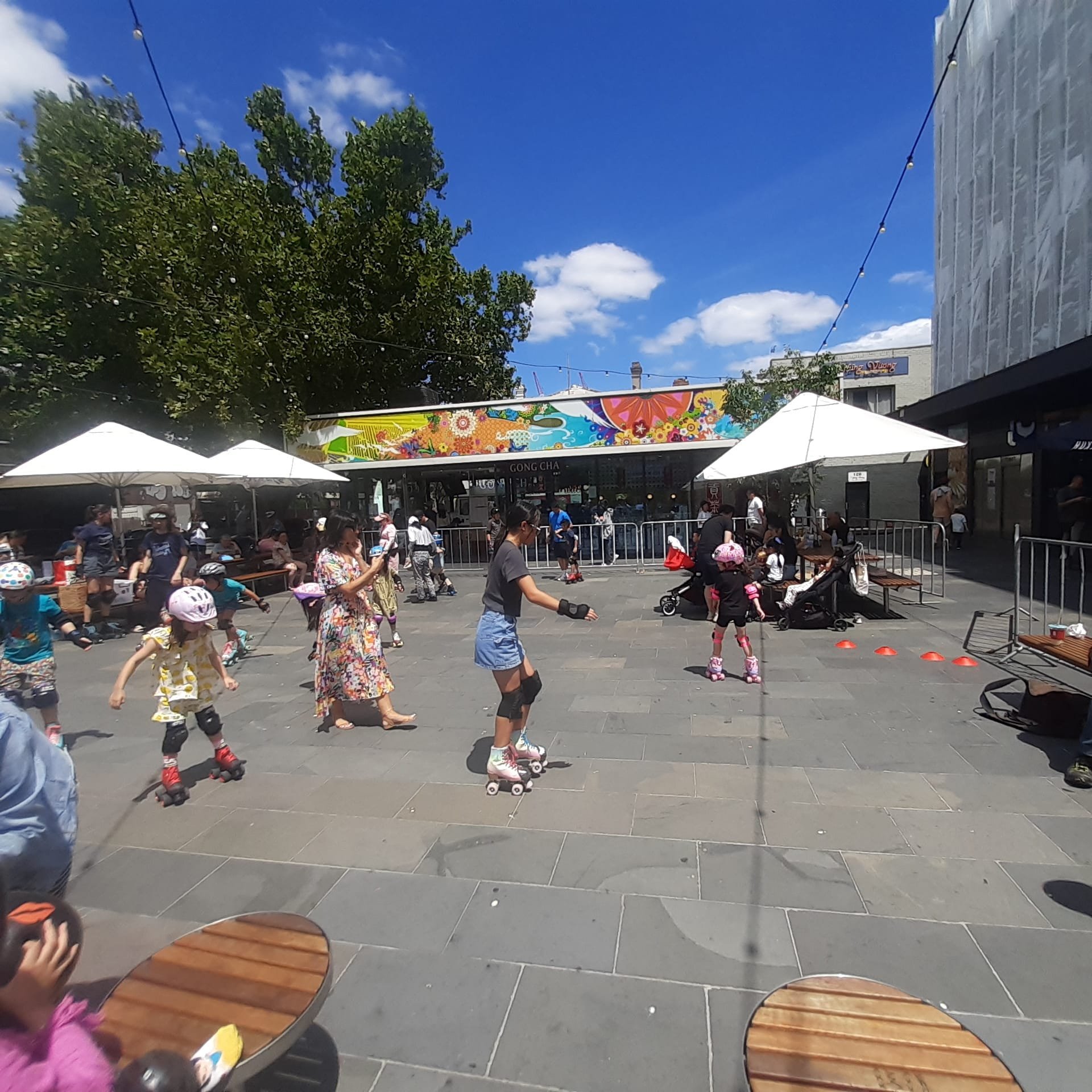 Children and adults roller skating in an outdoor plaza with colorful decorations, people seated under umbrellas, and sunny weather.