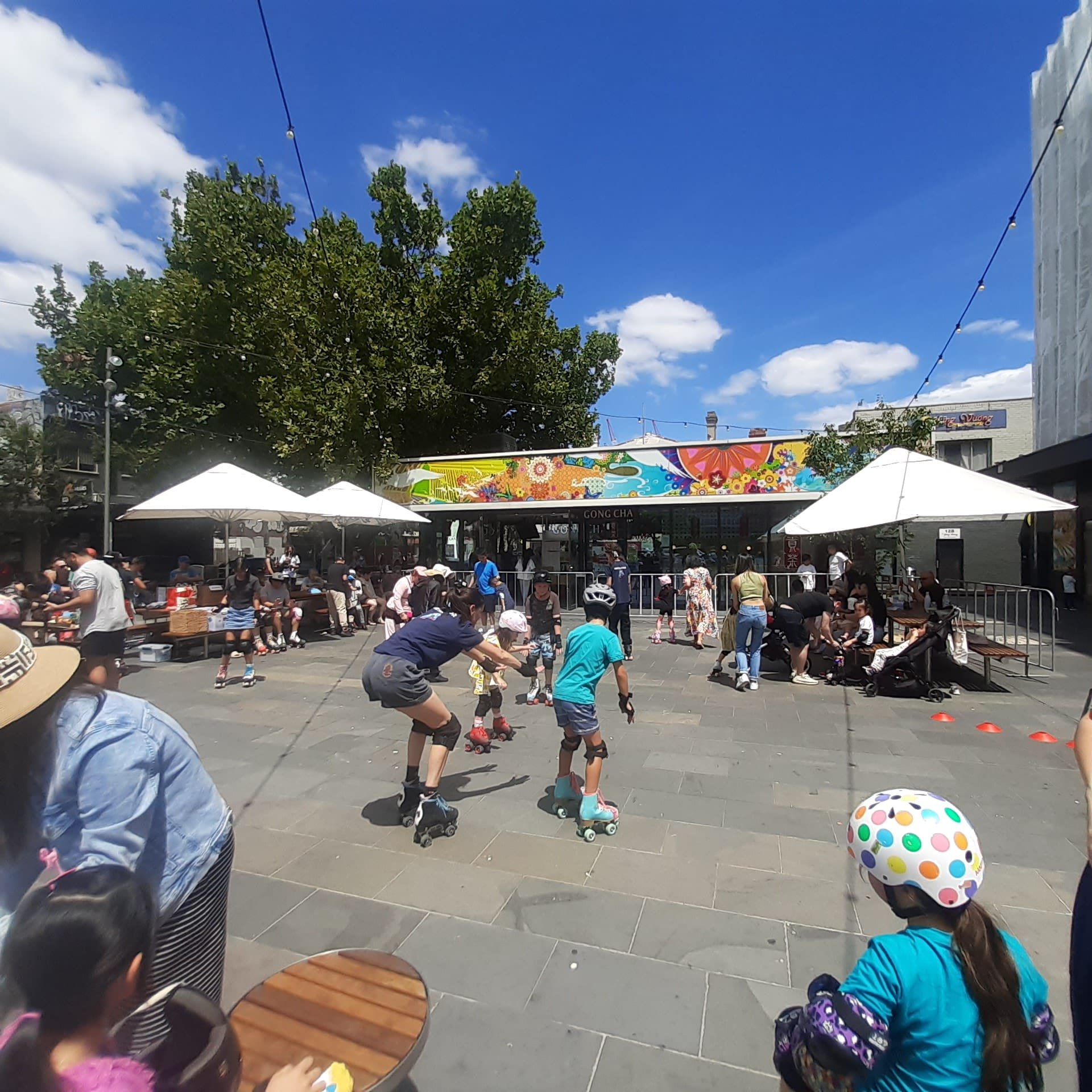 Outdoor event with people roller skating, colorful mural in background, white umbrellas, and trees.