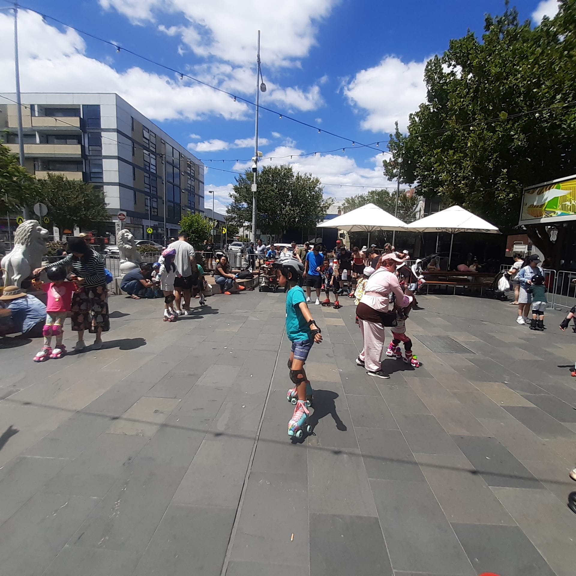 Outdoor scene with people roller skating in a plaza under a sunny sky. Buildings, trees, and a seating area with umbrellas are visible in the background.
